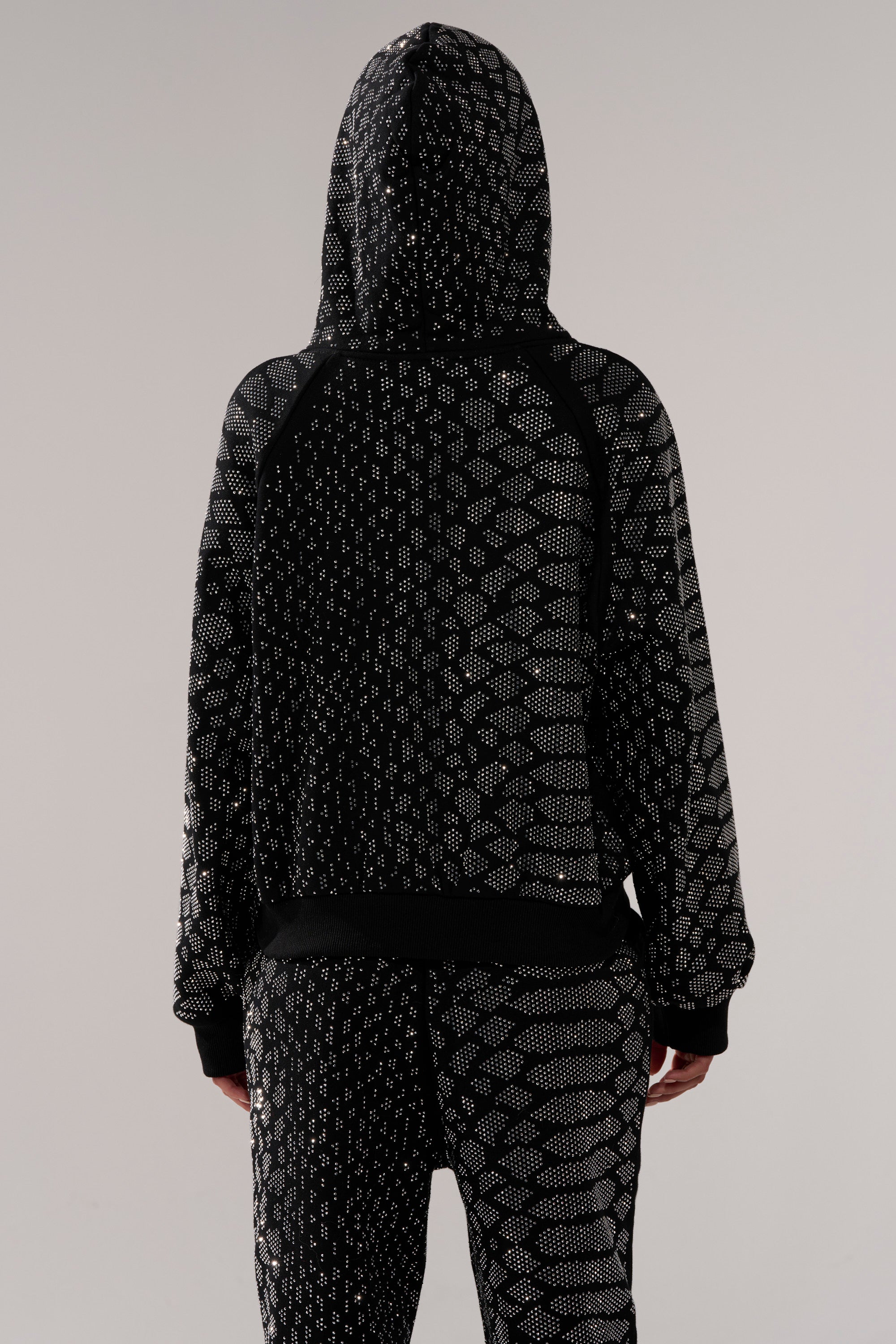 Someone stands facing away from the camera against a neutral background, wearing the SNEAKY LITTLE SNAKE ZIP UP SWEATSHIRT and matching pants featuring black fabric with a silver dotted geometric pattern.