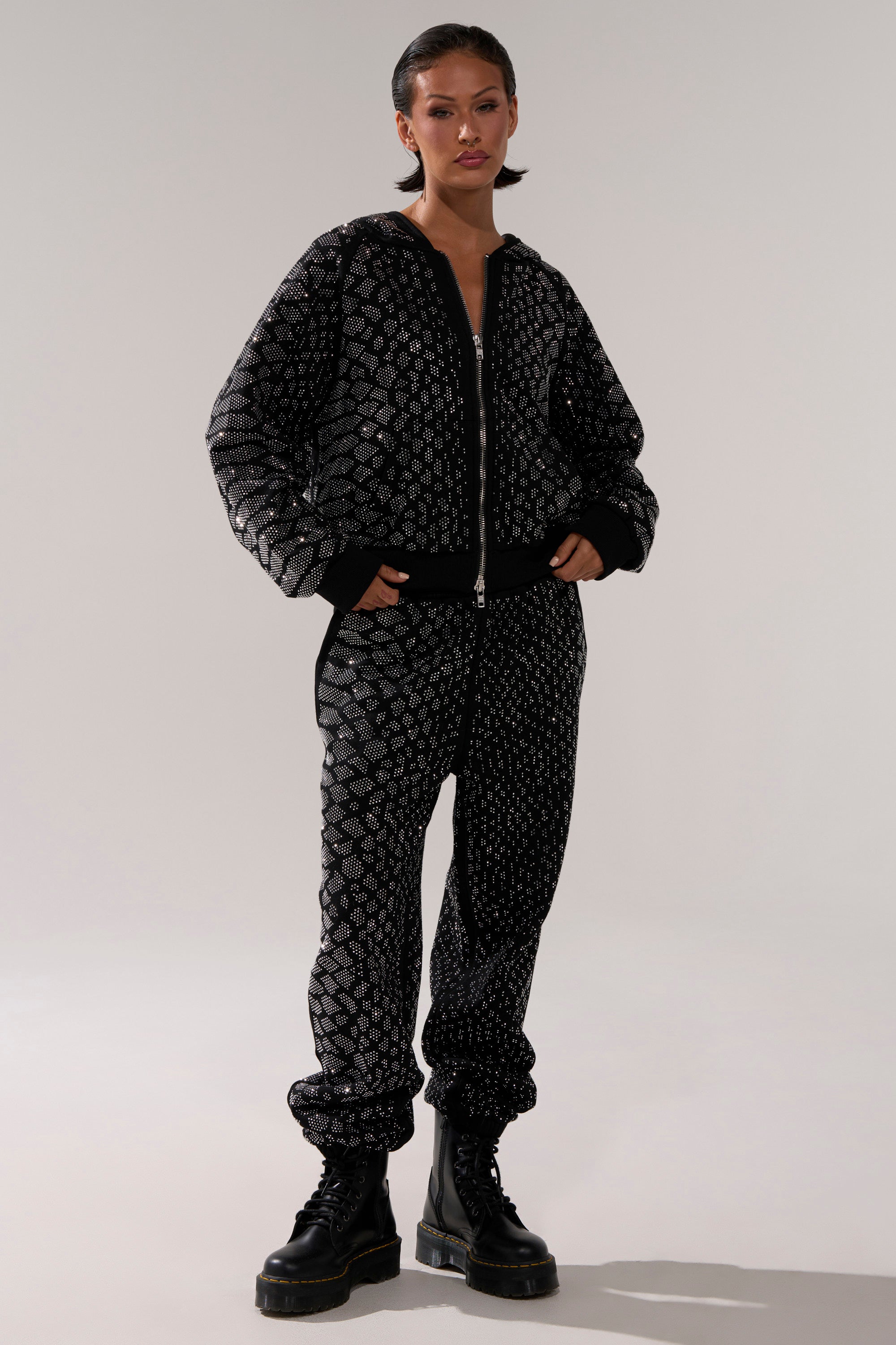 A woman stands confidently with hands in pockets, wearing the SNEAKY LITTLE SNAKE ZIP UP SWEATSHIRT and matching silver geometric pants, paired with black chunky boots and slicked-back hair.