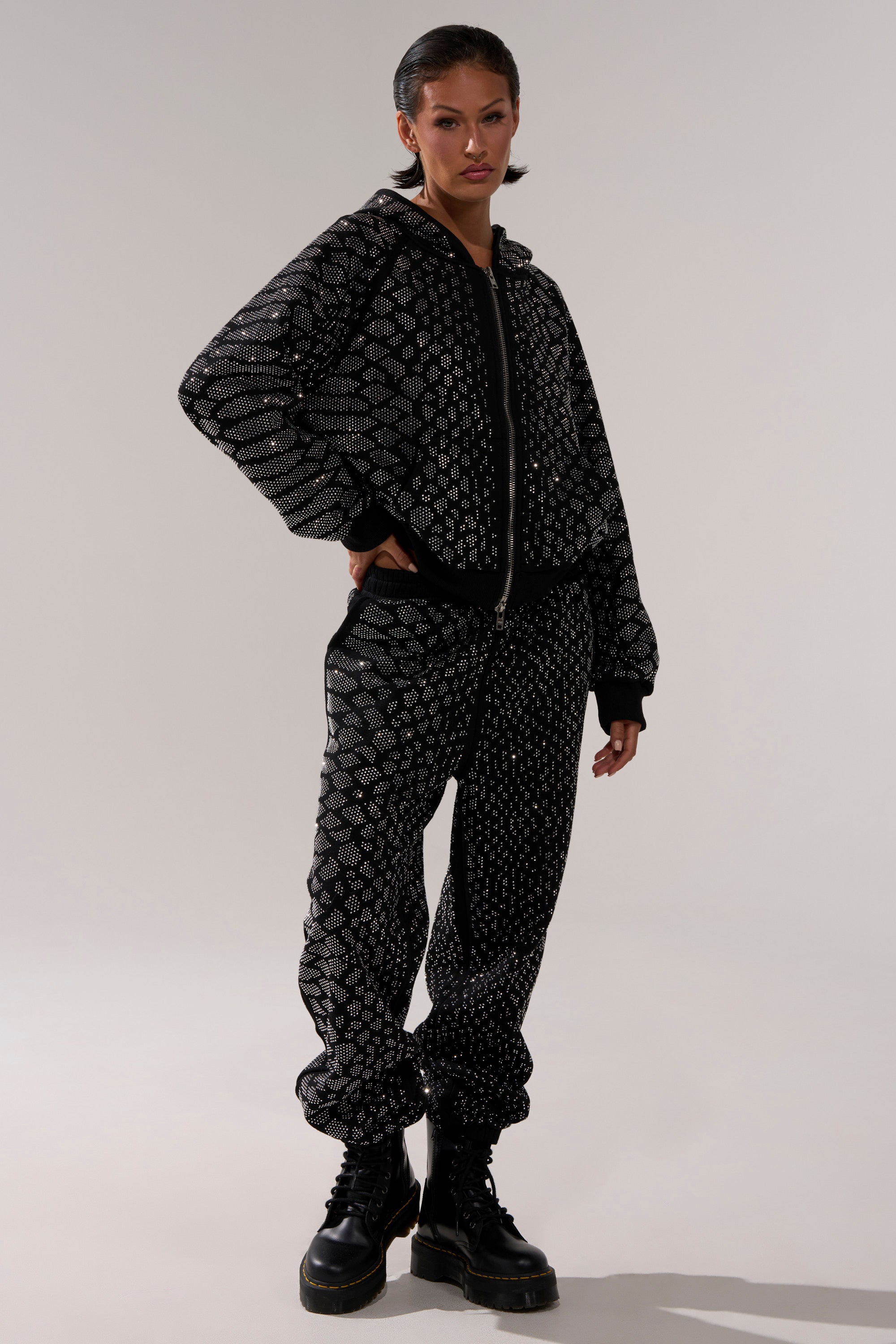A person stands confidently in the SNEAKY LITTLE SNAKE ZIP UP SWEATSHIRT, a black tracksuit with a white geometric pattern, paired with black lace-up boots against a plain light background.