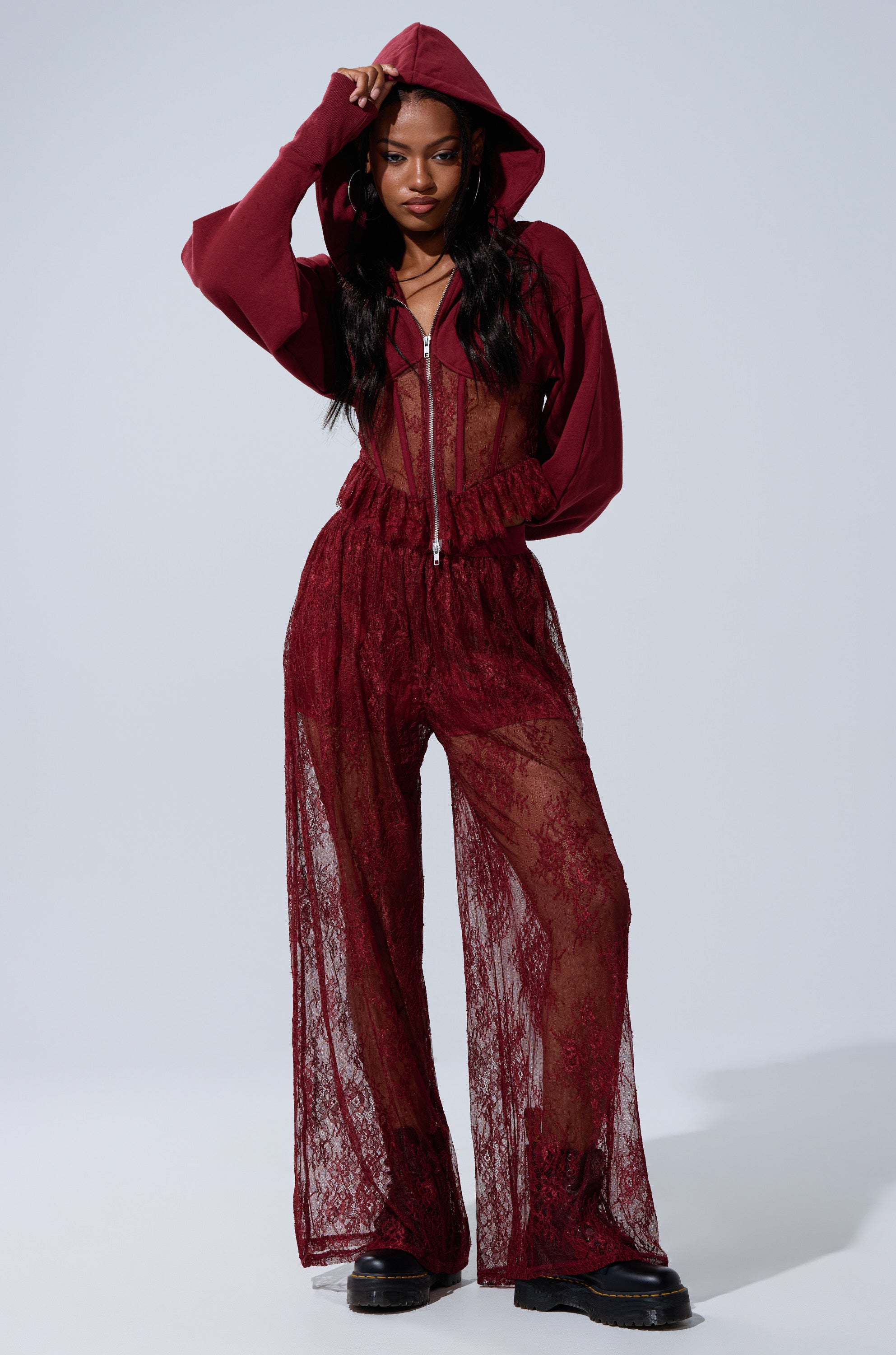 A woman models the TOO TRENDY LACE PANTS, a sheer burgundy lace wide-leg style with a matching hoodie. She pairs them with black platform shoes and showcases the bikini-style underlayer against a plain background. 0