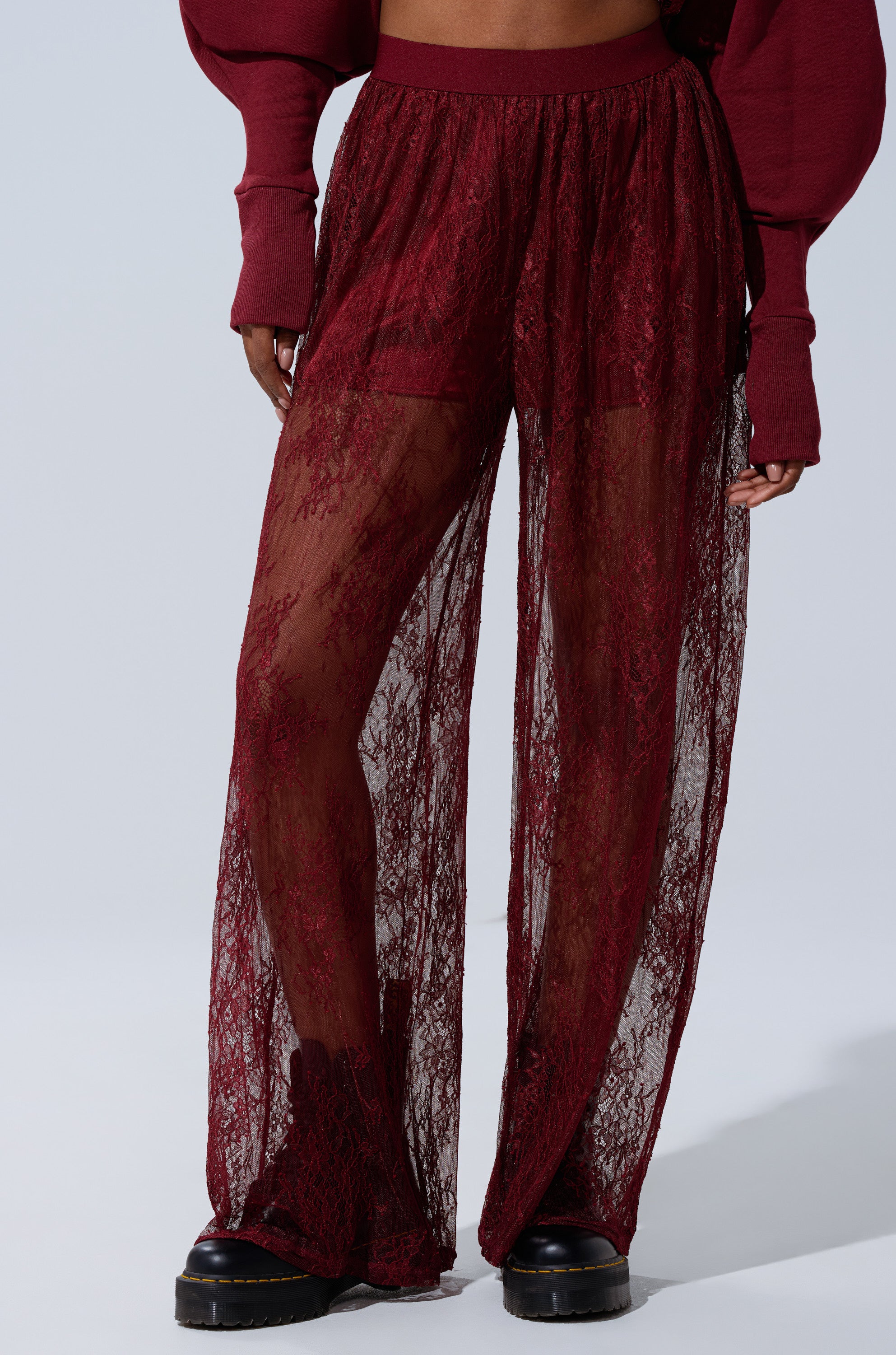 A person wears TOO TRENDY LACE PANTS in sheer burgundy lace with wide legs, revealing shorts underneath, paired with black platform shoes and a matching burgundy top. 1