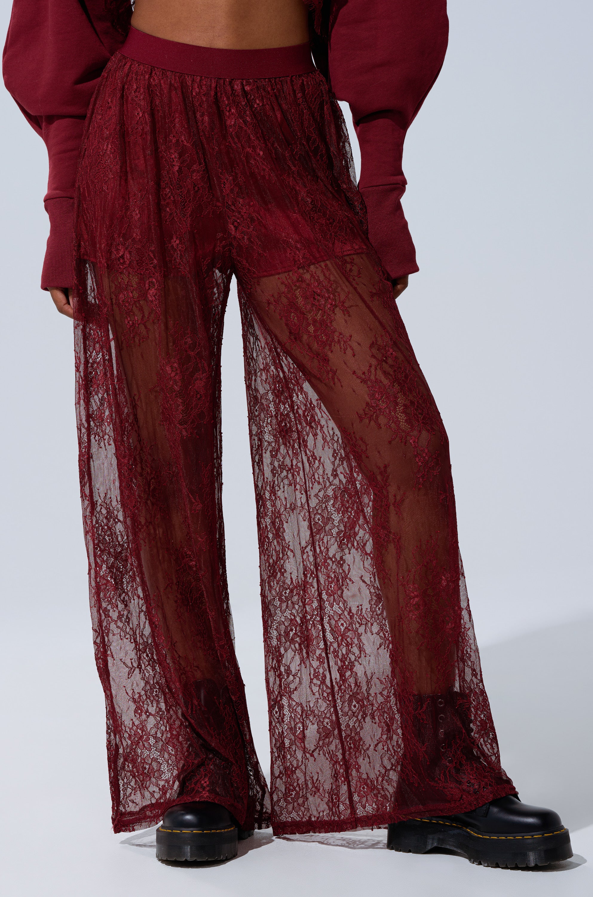 A person sports the TOO TRENDY LACE PANTS in sheer burgundy over matching shorts, styled with chunky black boots and a burgundy long-sleeve top. The bold, wide-leg lace pants make a striking statement in this standout look. 2