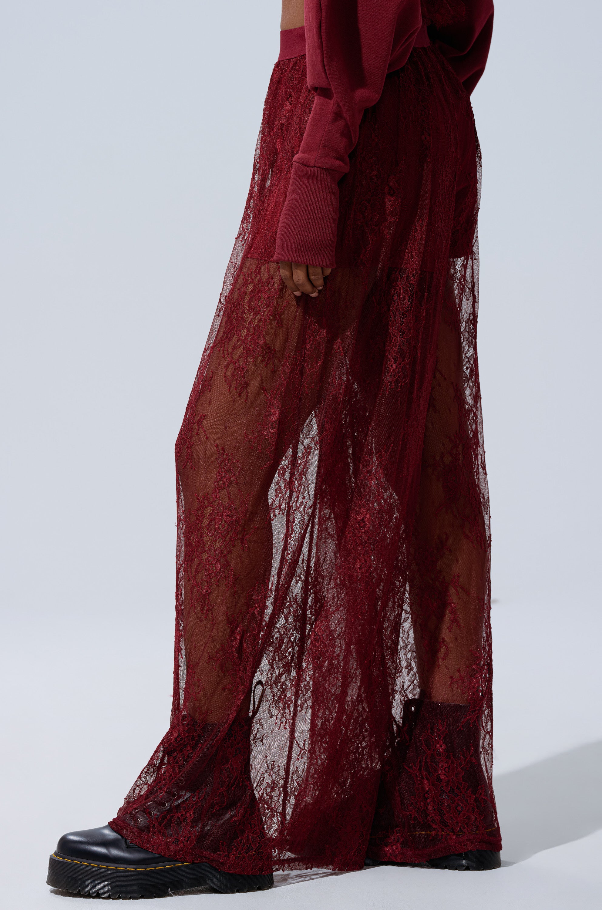 A person models the TOO TRENDY LACE PANTS—long, sheer burgundy lace with a high-rise waist over matching shorts—styled with a burgundy top and black chunky heels, against a plain light background. 3