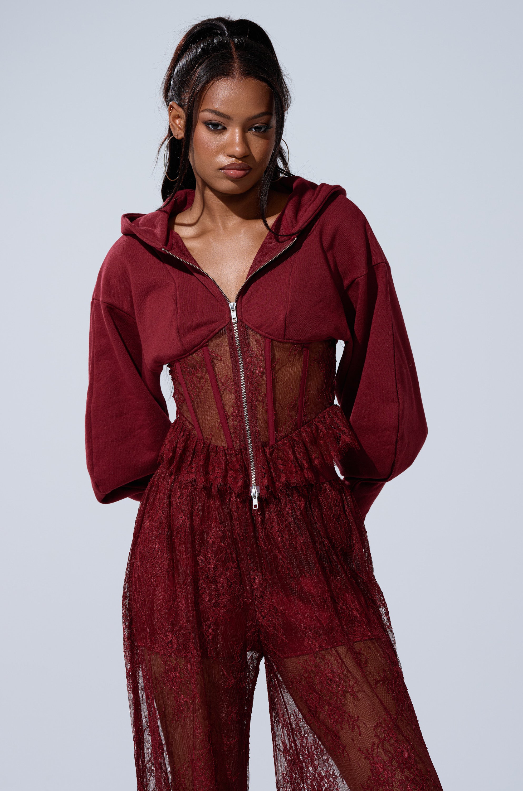 A woman stands against a light gray background, confidently wearing the TOO TRENDY ZIP UP SWEATSHIRT WITH LACE over a sheer burgundy lace top and matching high-waisted lace pants.