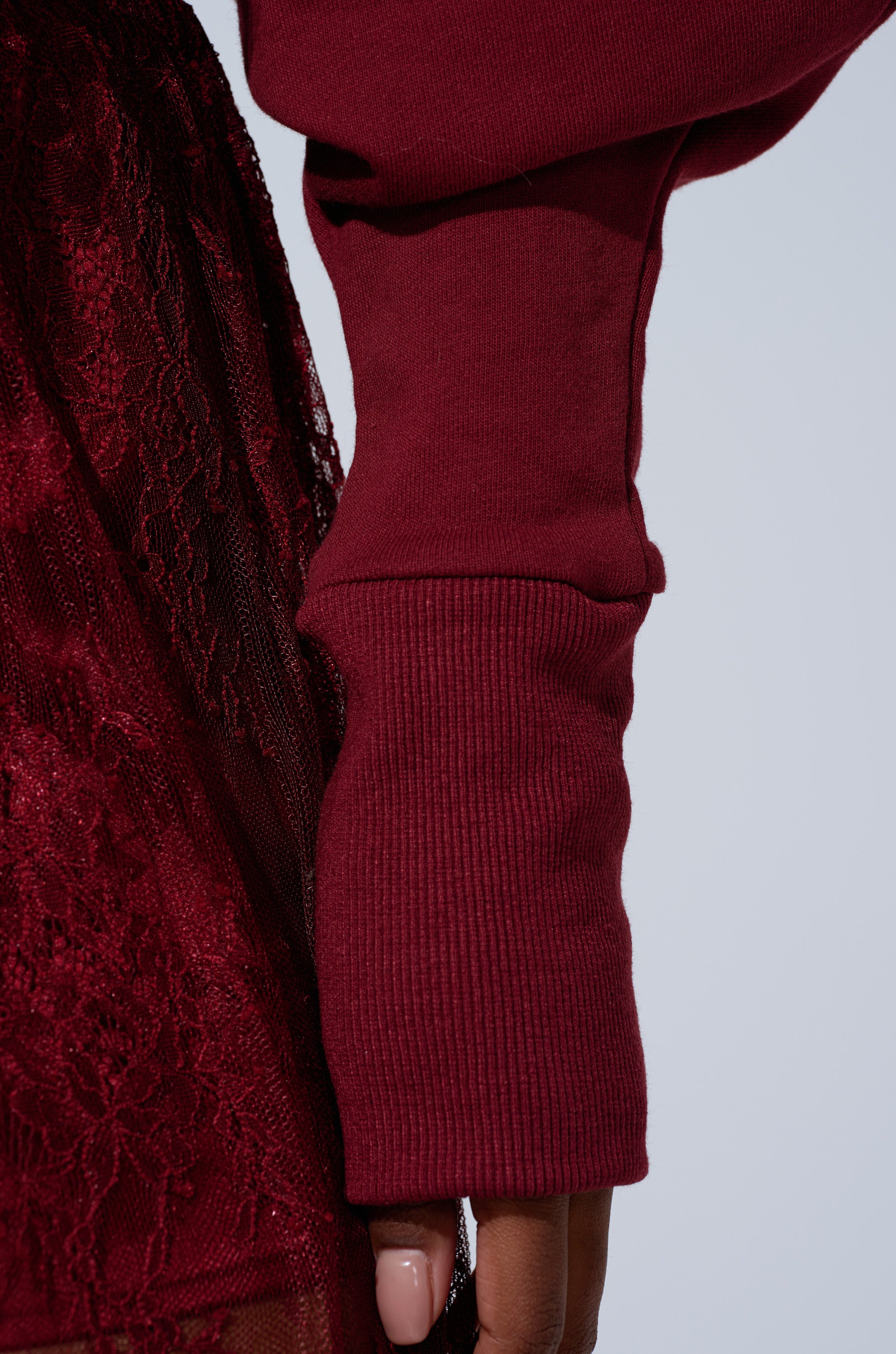 A close-up of a person wearing the TOO TRENDY ZIP UP SWEATSHIRT WITH LACE in burgundy, styled with a matching lace skirt; their hand is partially visible with nude polished nails.