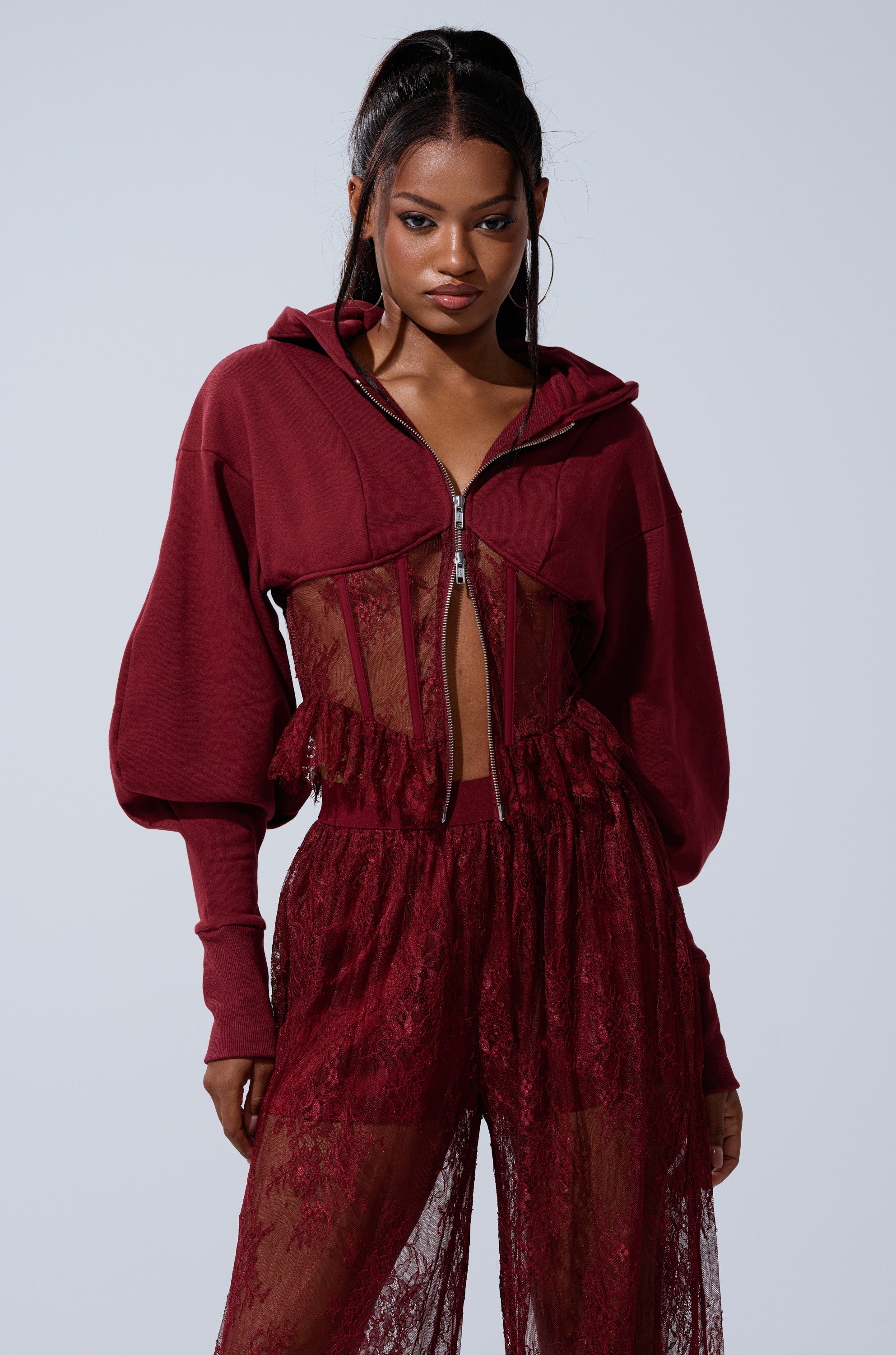 A woman confidently models the TOO TRENDY ZIP UP SWEATSHIRT WITH LACE in burgundy, pairing the lace hoodie with matching sheer lace pants against a simple light background.