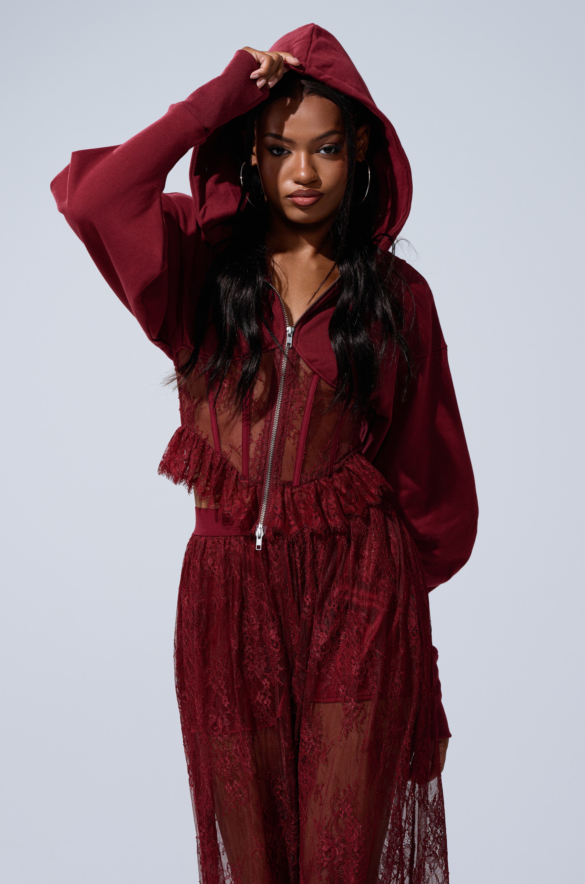 A woman models the TOO TRENDY ZIP UP SWEATSHIRT WITH LACE in burgundy, pairing it with a sheer lace dress. She holds the hood with one hand and gazes confidently at the camera against a plain light background.