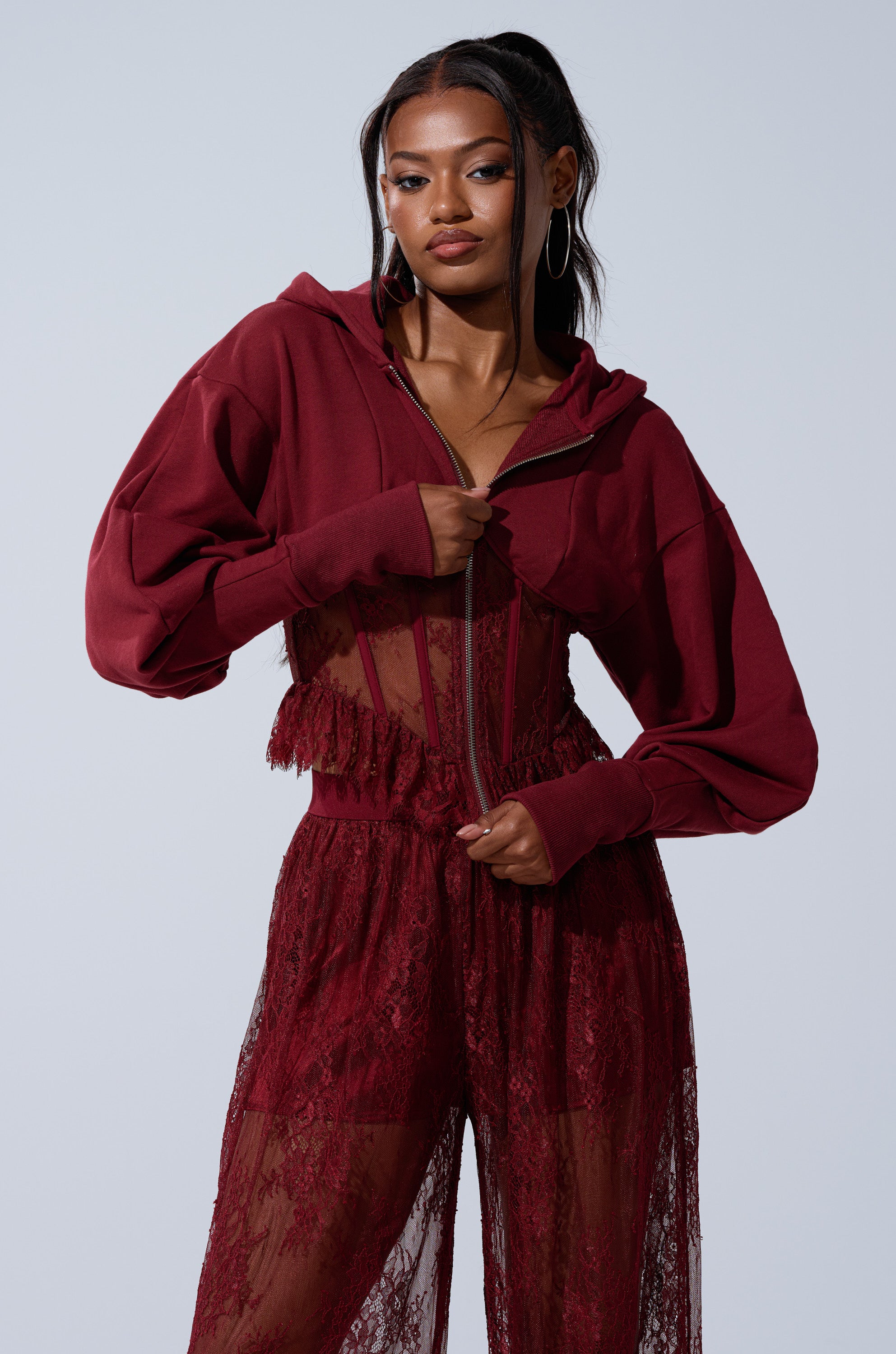 A woman poses confidently against a light gray background, wearing the TOO TRENDY ZIP UP SWEATSHIRT WITH LACE in burgundy over a matching sheer lace jumpsuit.