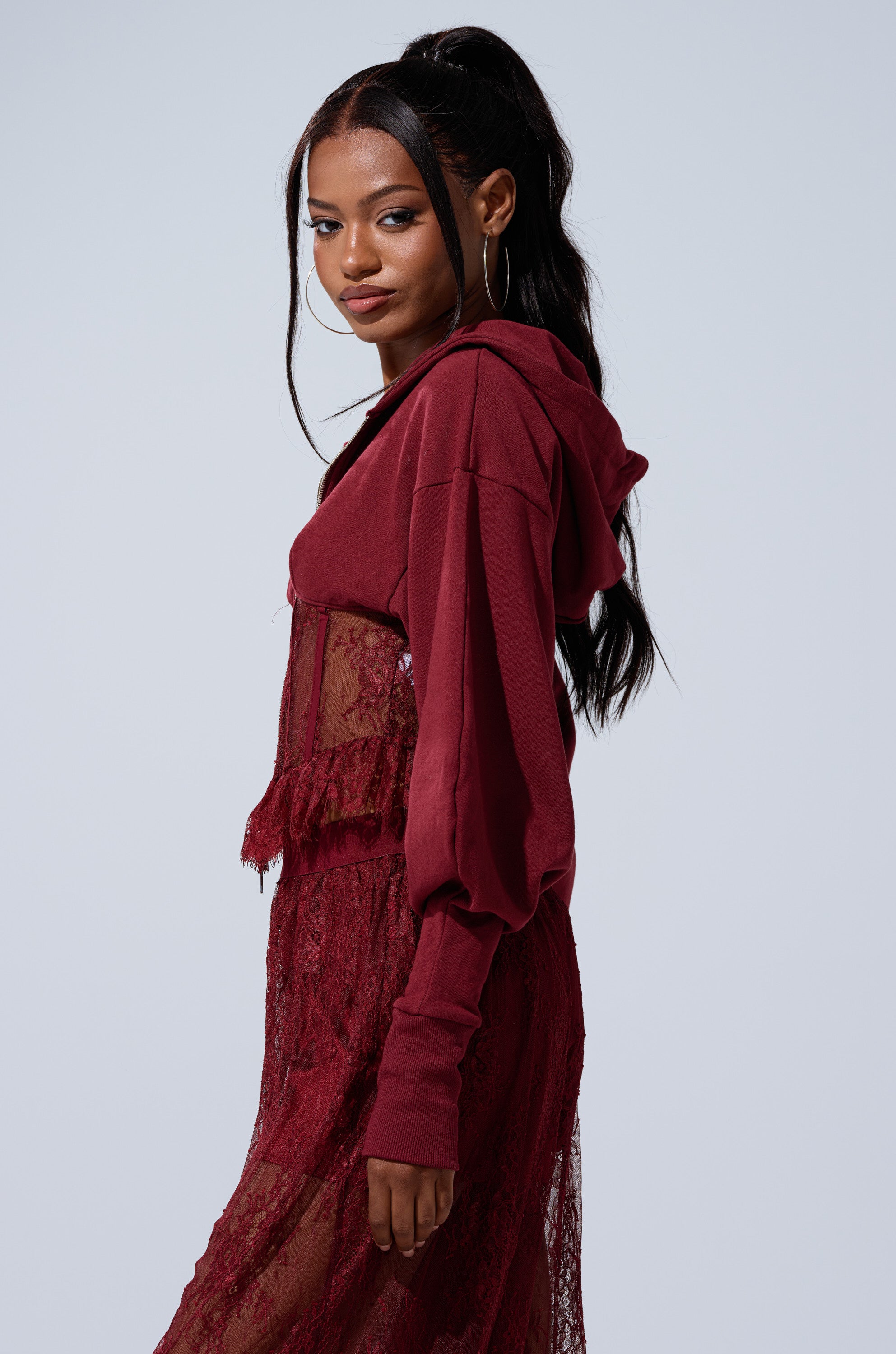 A woman stands sideways, smiling at the camera, wearing the TOO TRENDY ZIP UP SWEATSHIRT WITH LACE layered over a sheer maroon lace dress. Her long dark hair is styled in a high ponytail.