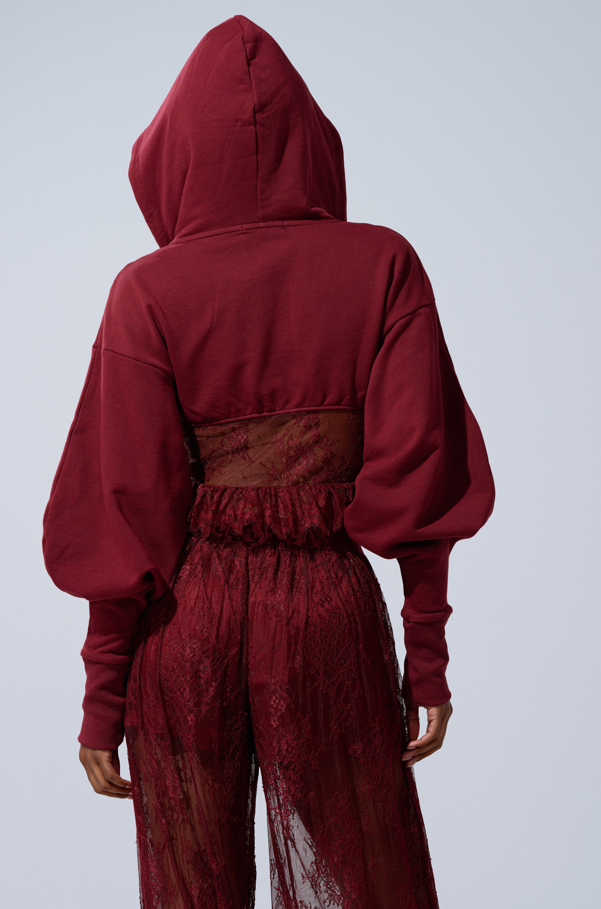 A person stands with their back turned, wearing the TOO TRENDY ZIP UP SWEATSHIRT WITH LACE—a cropped burgundy hoodie with voluminous sleeves and sheer, high-waisted lace pants. The hood is up, covering their head.