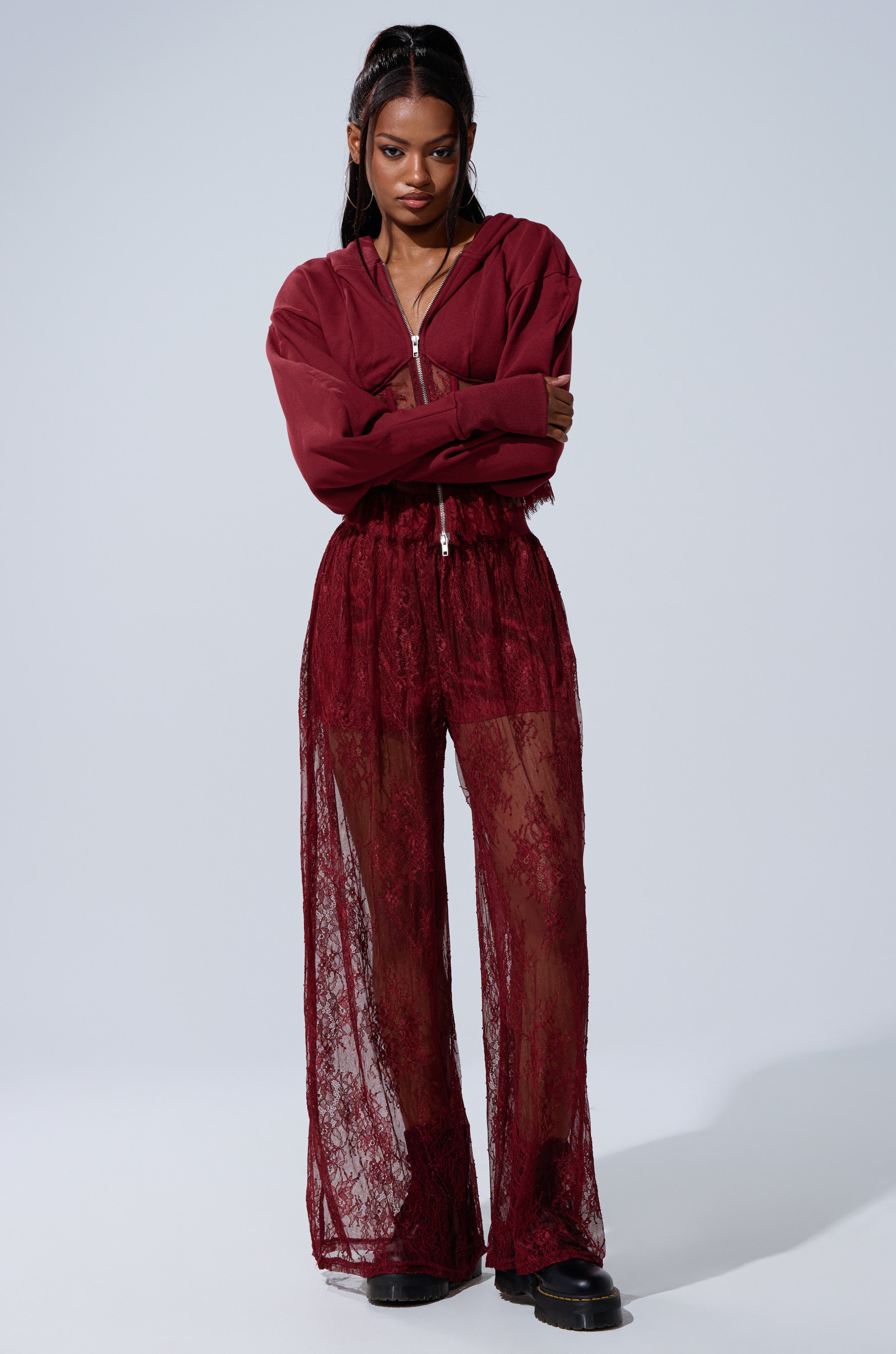 A woman stands with arms crossed, wearing the TOO TRENDY ZIP UP SWEATSHIRT WITH LACE, paired with sheer burgundy lace pants over black shorts. Her long dark hair is in half-up pigtails, and she finishes the outfit with black platform shoes.