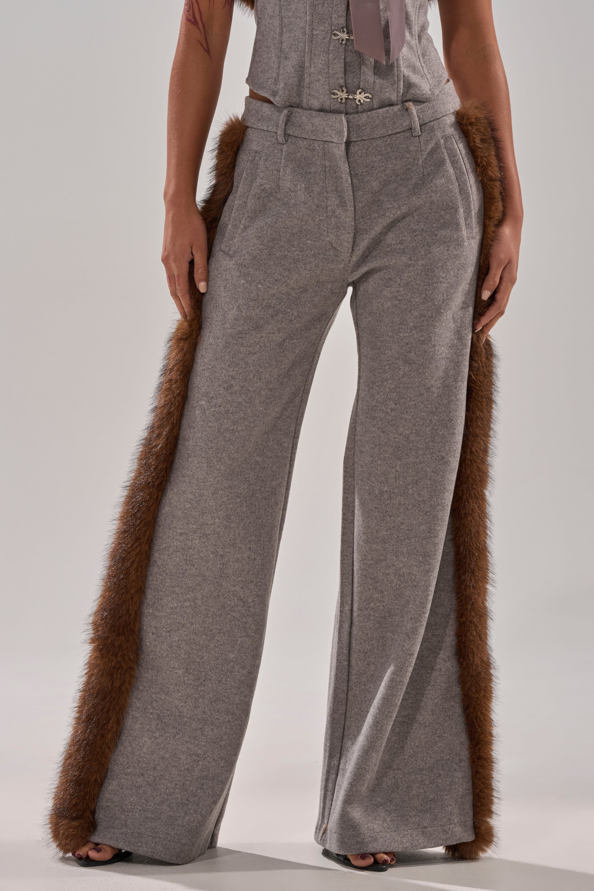 A person wears the HALLE FUR TRIM WIDE LEG PANT, featuring wool-like fabric and brown faux fur trim along the sides, styled with a matching gray top for a sleek, modern look.