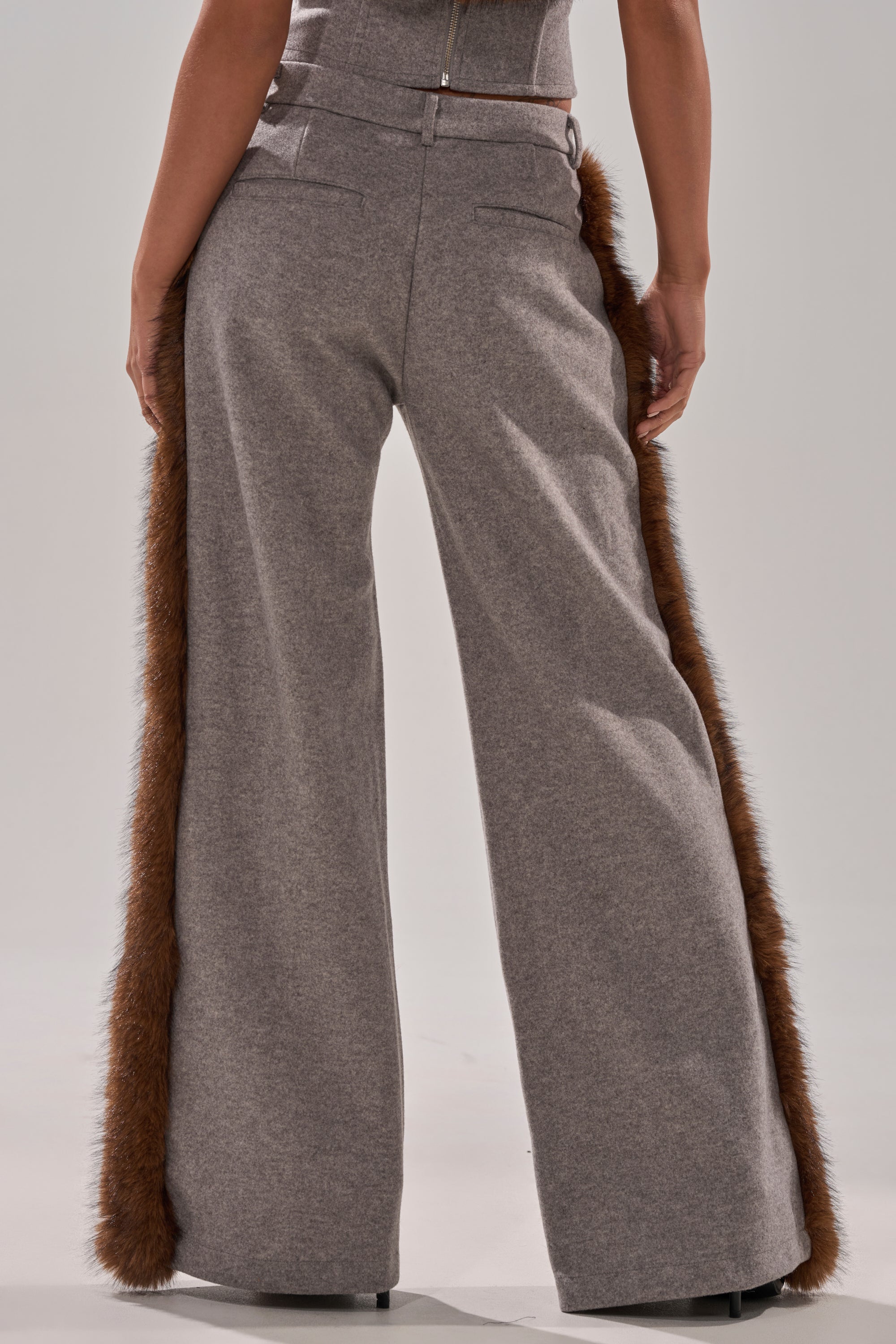 A person wearing the HALLE FUR TRIM WIDE LEG PANT, featuring high-waisted wide legs and brown faux fur trim along the outer seams, stands with their back to the camera.