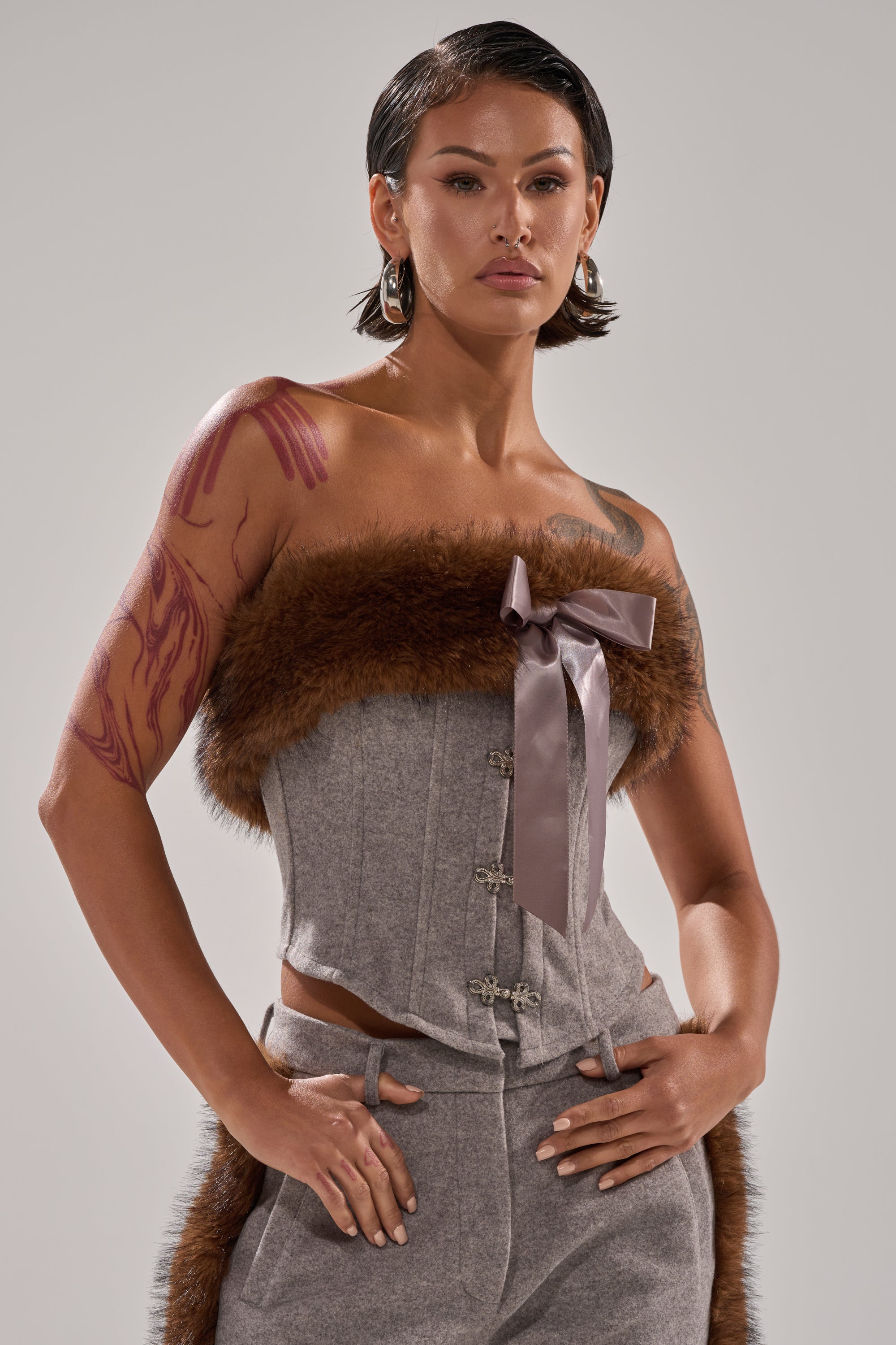 A person with short dark hair and hoop earrings confidently wears the HALLE FUR TRIM CORSET in gray with brown faux fur and a large satin bow, paired with matching pants; their arm tattoos are visible.