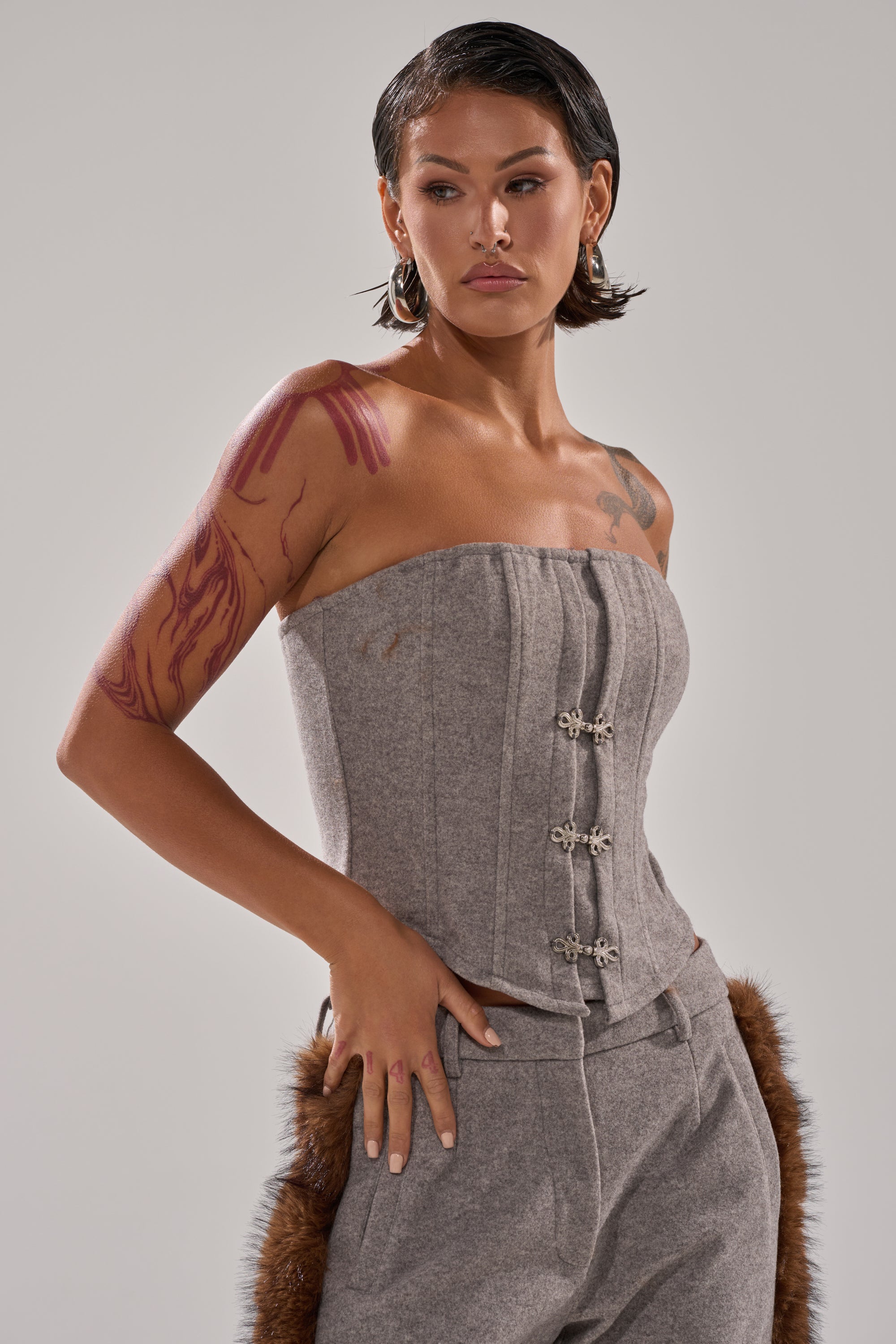 A woman with short dark hair and visible tattoos wears the HALLE FUR TRIM CORSET with matching pants, standing against a plain light background.