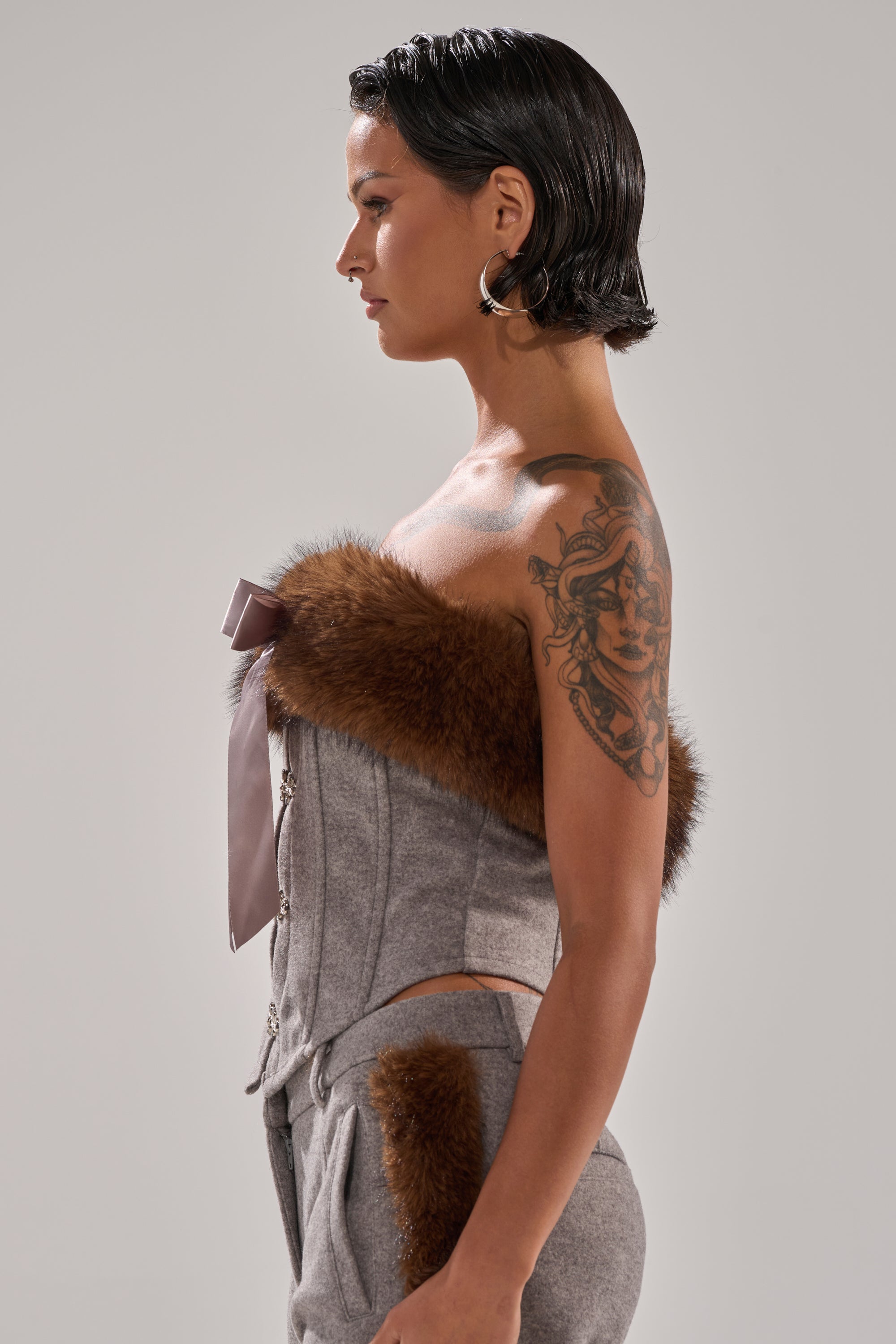 A person with short dark hair and a large arm tattoo poses in profile, wearing the HALLE FUR TRIM CORSET with a strapless neckline and matching wool-like pants, set against a plain light background.