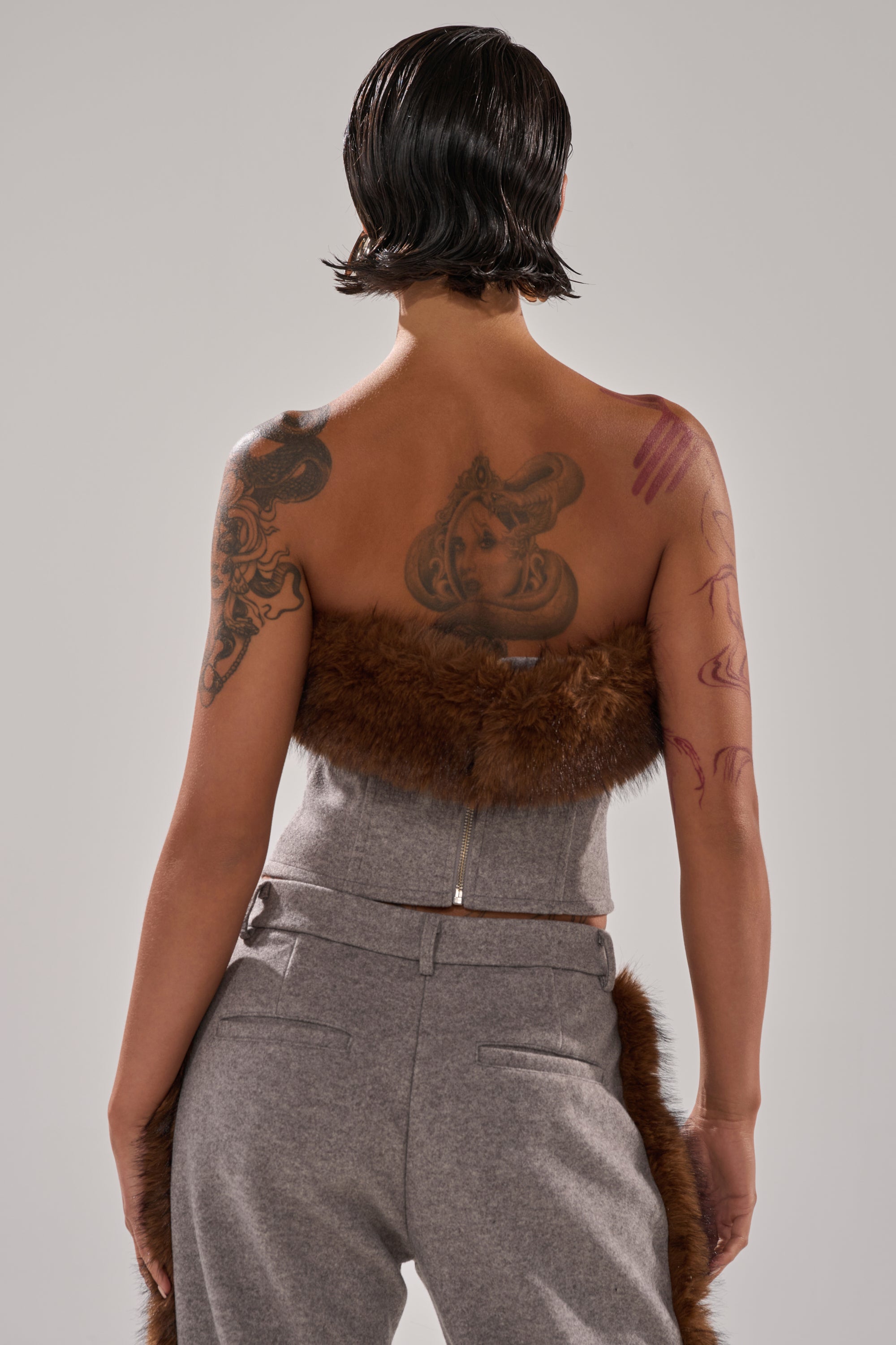 A person with short, wet-look dark hair is seen from behind, wearing gray pants and the HALLE FUR TRIM CORSET with a strapless neckline. Their back and arms showcase several tattoos, including a large upper back design.