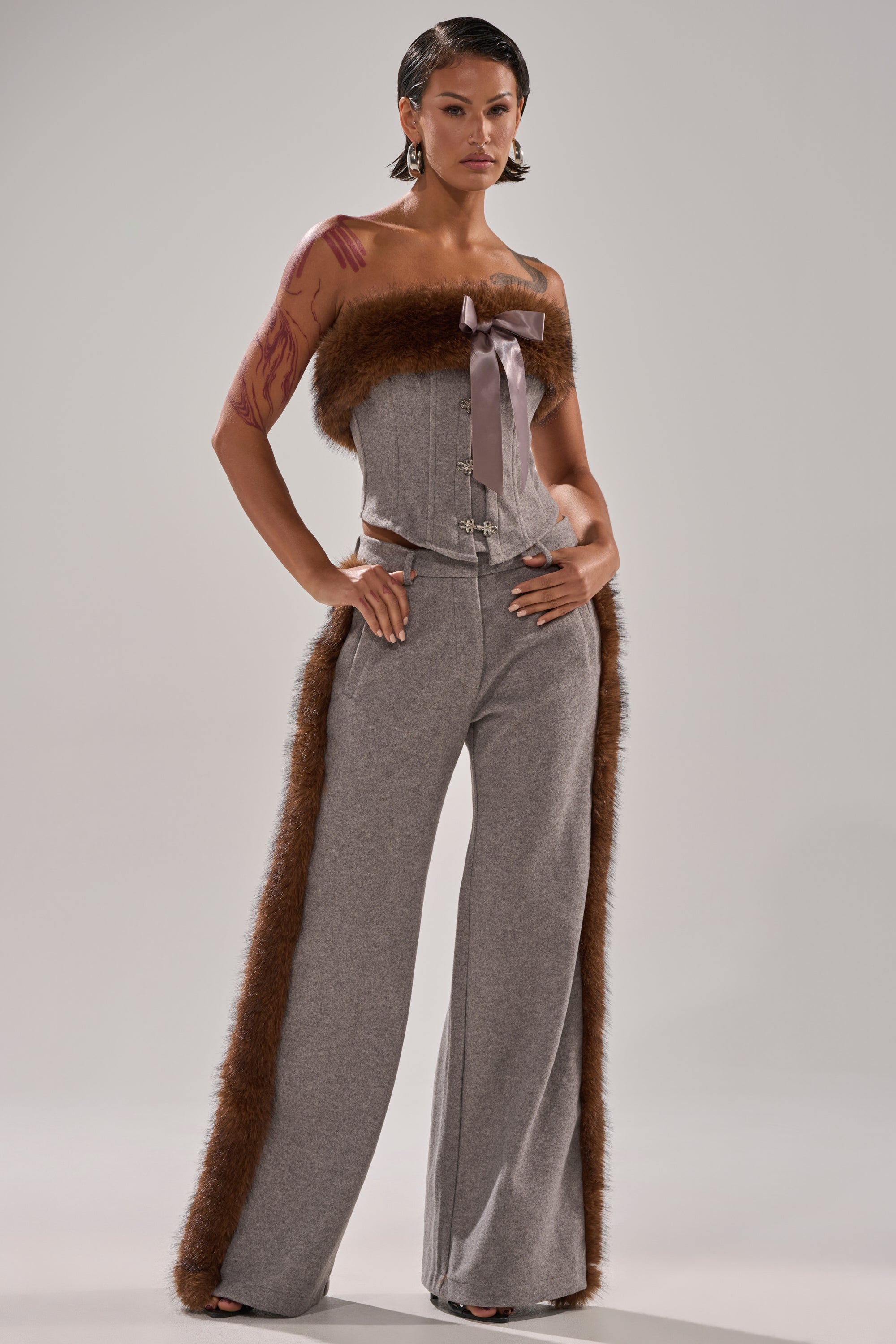 A model wears the HALLE FUR TRIM CORSET, a strapless gray jumpsuit with brown faux fur trim along the neckline and down the sides of its wide-leg pants, standing confidently against a plain backdrop with short wet-look hair.