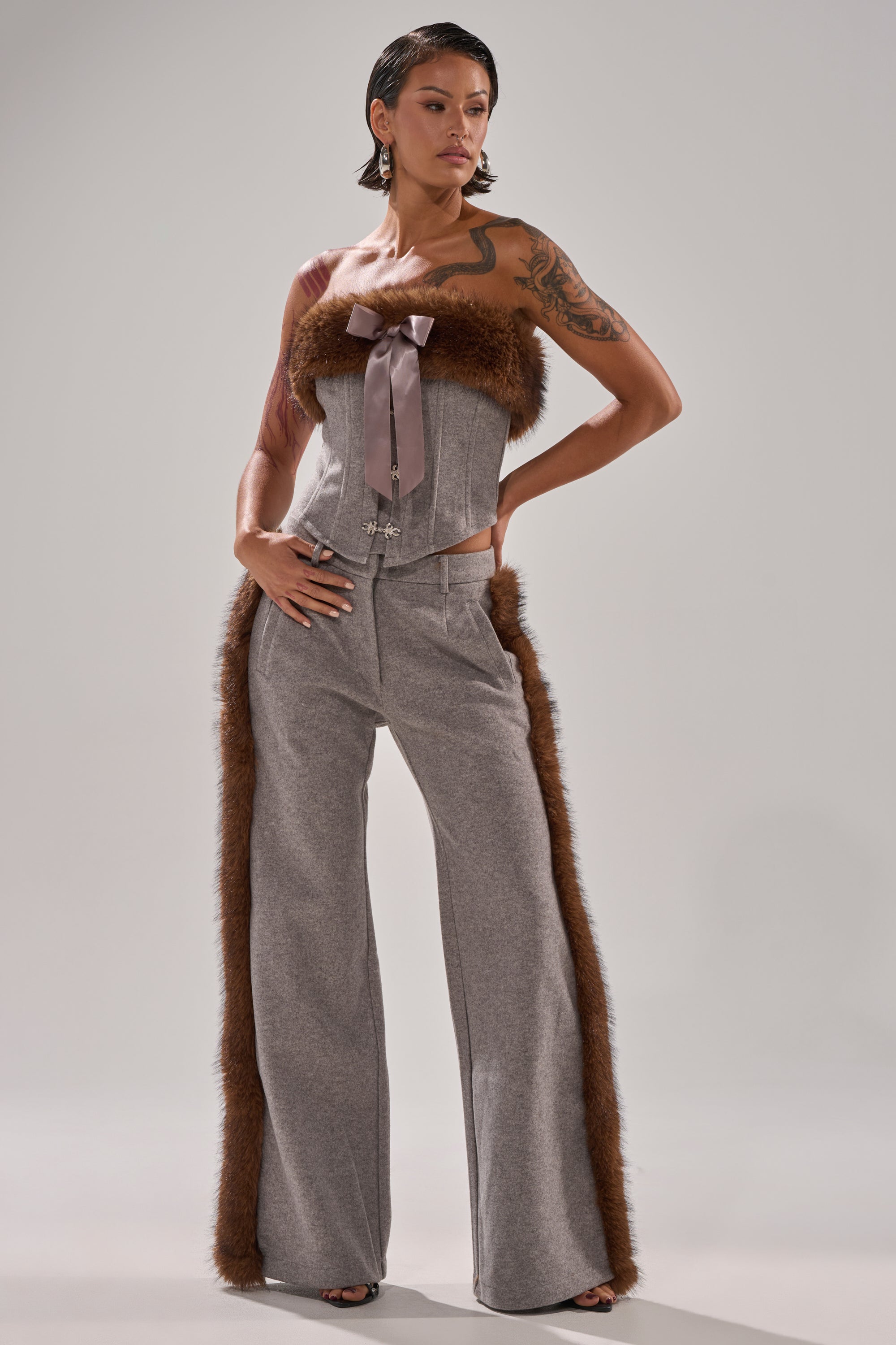 Wearing the HALLE FUR TRIM CORSET—a strapless gray top with brown fur and a wide ribbon—paired with matching gray pants featuring brown fur sides, a person stands confidently against a plain light background.