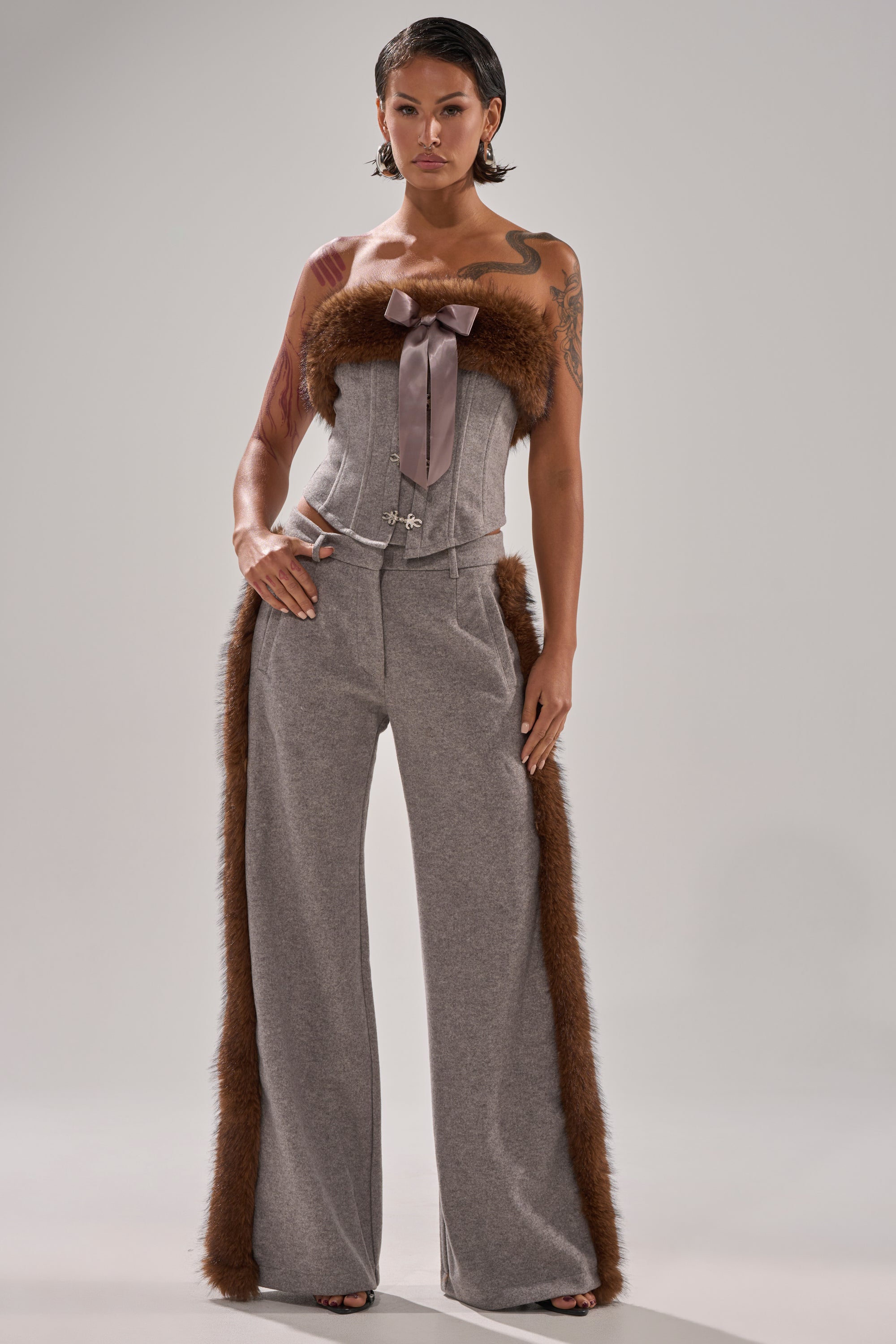 A woman with short dark hair and visible tattoos wears the HALLE FUR TRIM CORSET—a gray strapless top with a pink ribbon and brown fur trim—paired with matching wide-leg gray pants featuring brown fur along the sides.