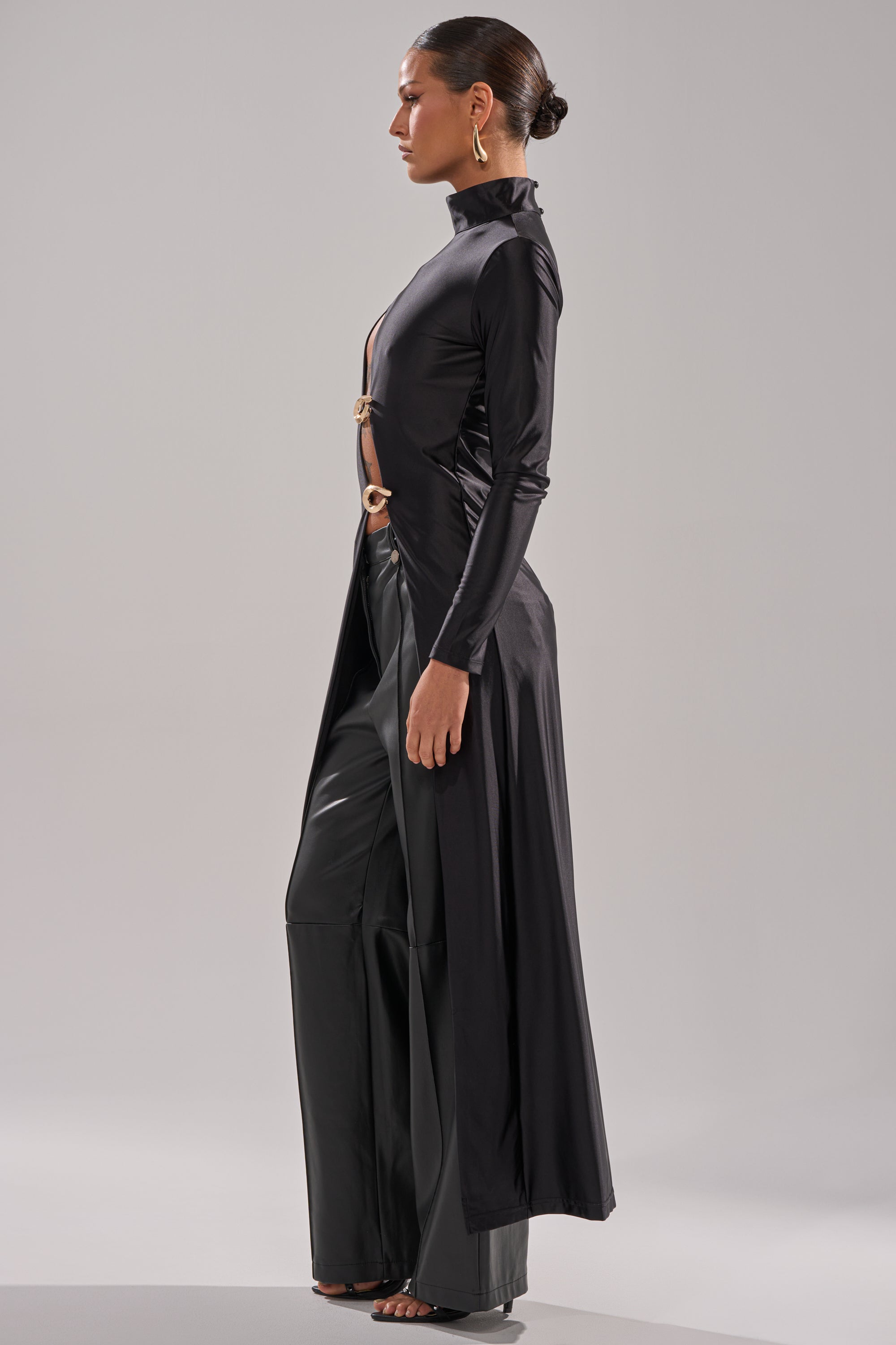 A woman stands in profile wearing the ITS TIME TO CELEBRATE SATIN TURTLE NECK BLOUSE with side cutouts and gold ring details, paired with wide-leg black pants and large gold hoop earrings for an elegant evening look. 1