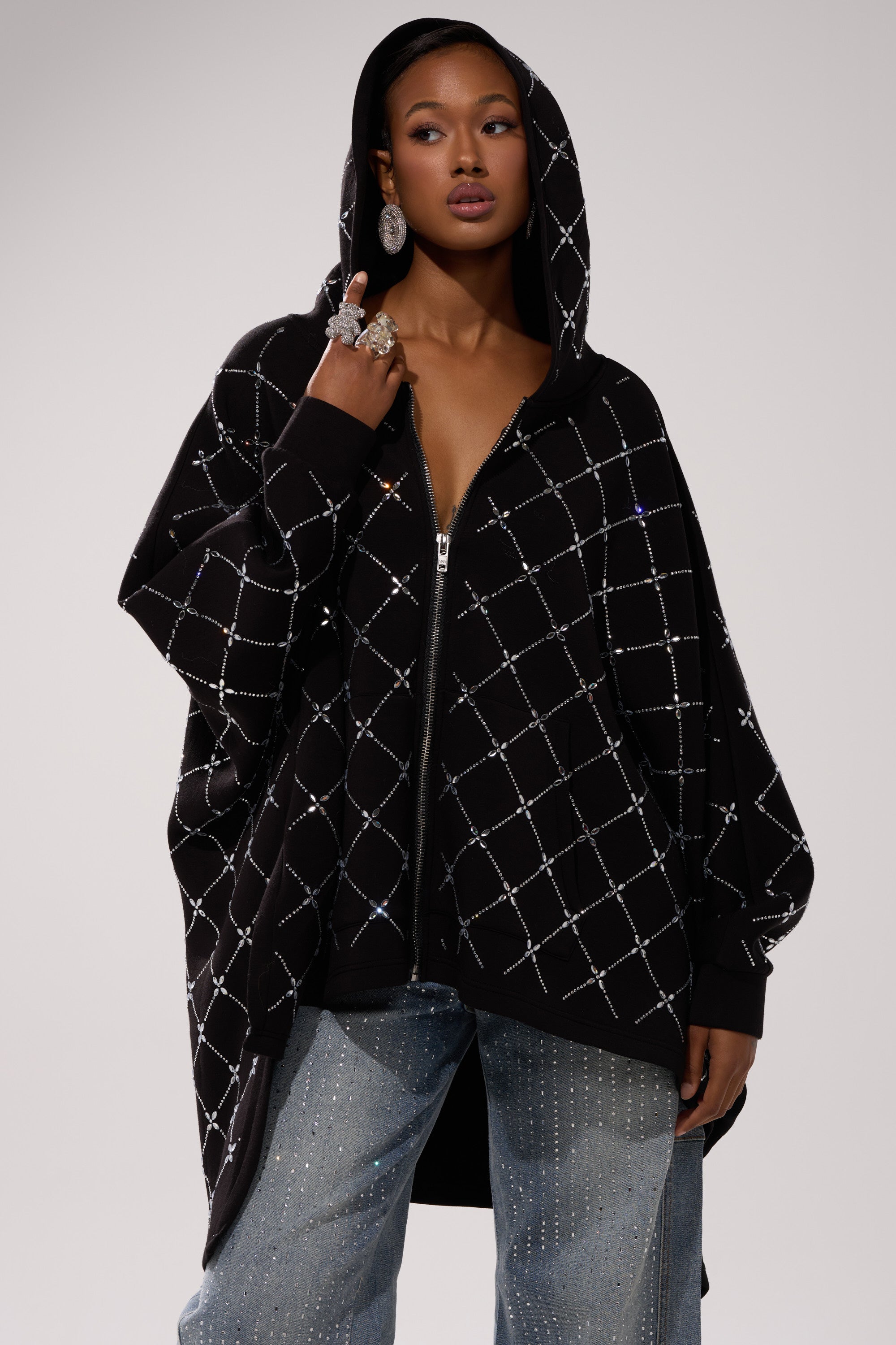 A woman poses confidently with the hood up, showcasing streetwear layering in the BLING BLING OVERSIZED ZIP UP SWEATSHIRT, paired with light blue rhinestone-detailed jeans and large statement earrings.