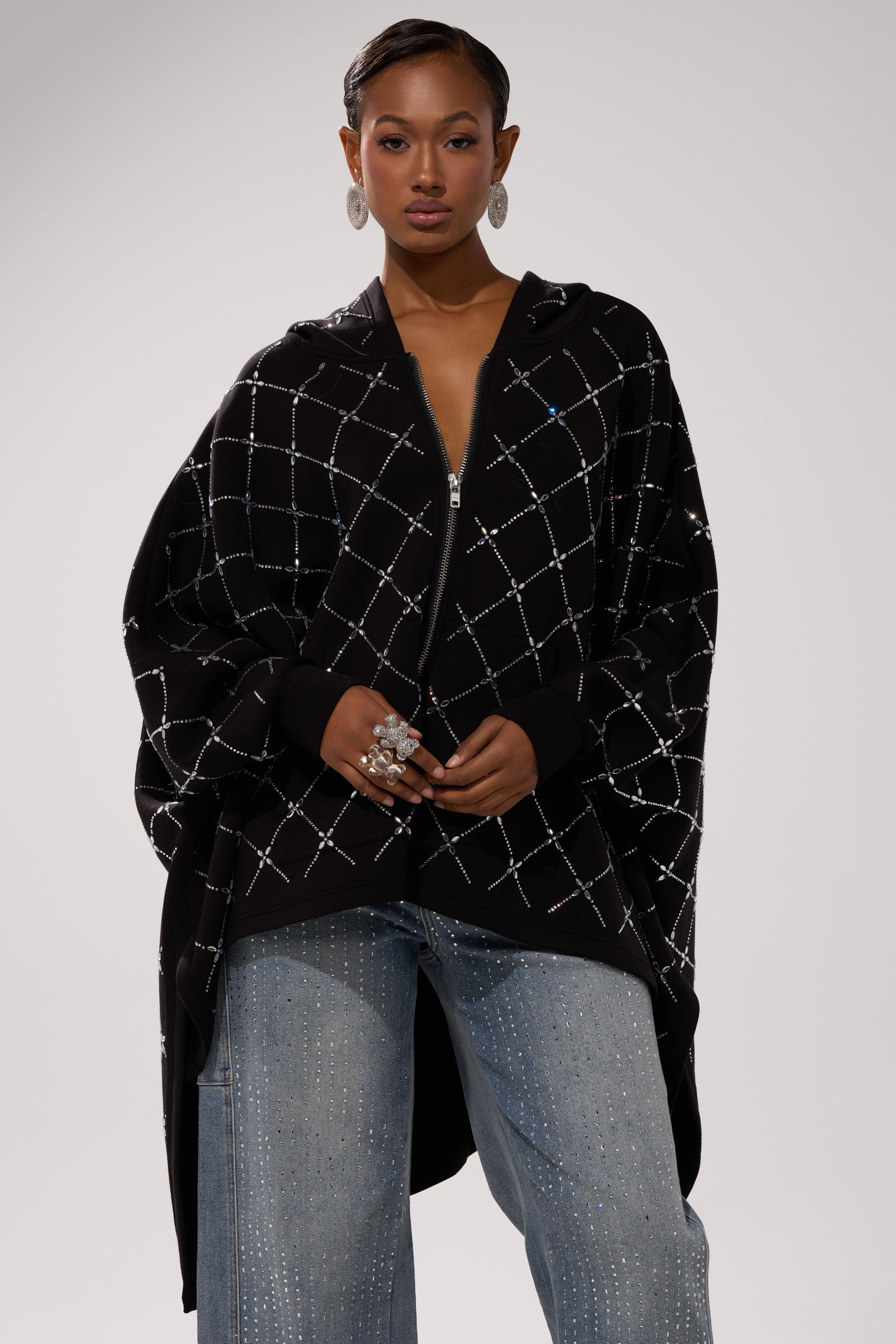 A woman faces forward in the BLING BLING OVERSIZED ZIP UP SWEATSHIRT with a white crisscross pattern, paired with light blue wide-leg sparkly jeans, bold rings, statement earrings, and slicked-back hair for a streetwear vibe.