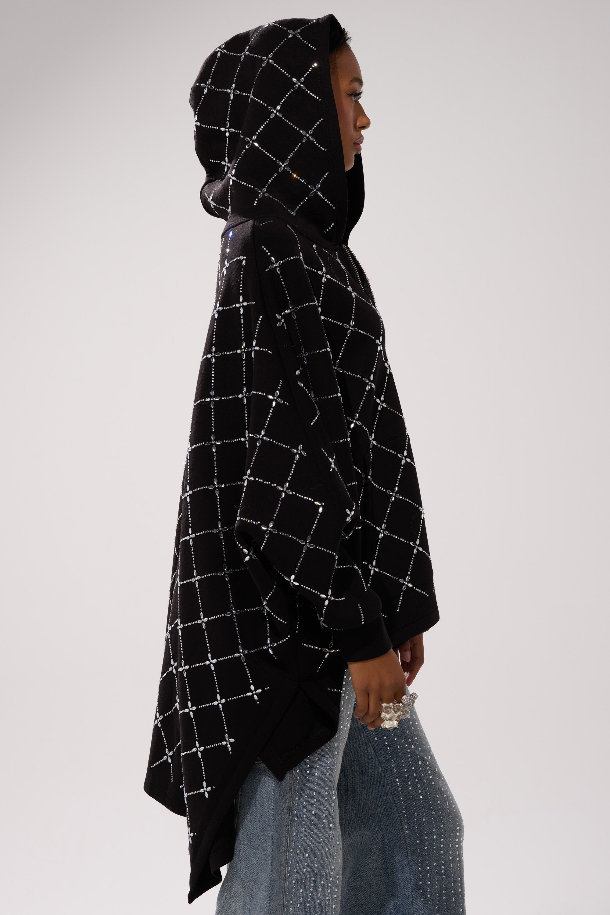 A person in profile wears the BLING BLING OVERSIZED ZIP UP SWEATSHIRT, a black streetwear-inspired hooded poncho with a white grid pattern, paired with blue jeans featuring silver rhinestone embellishments.