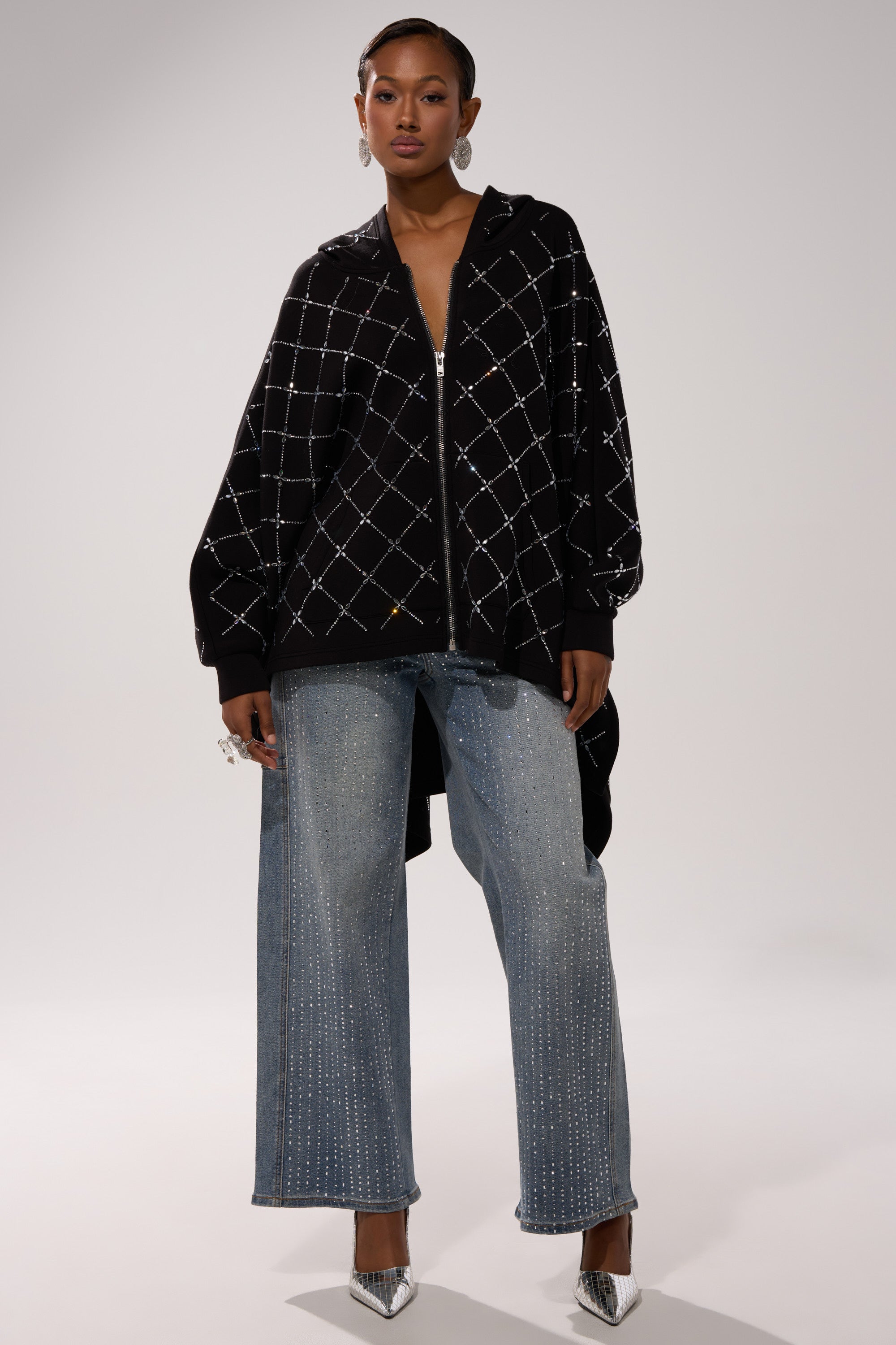 A woman faces forward wearing the BLING BLING OVERSIZED ZIP UP SWEATSHIRT with a black and white diamond pattern, wide-leg studded blue jeans, silver hoop earrings, and metallic pointed-toe heels—ideal for streetwear layering.