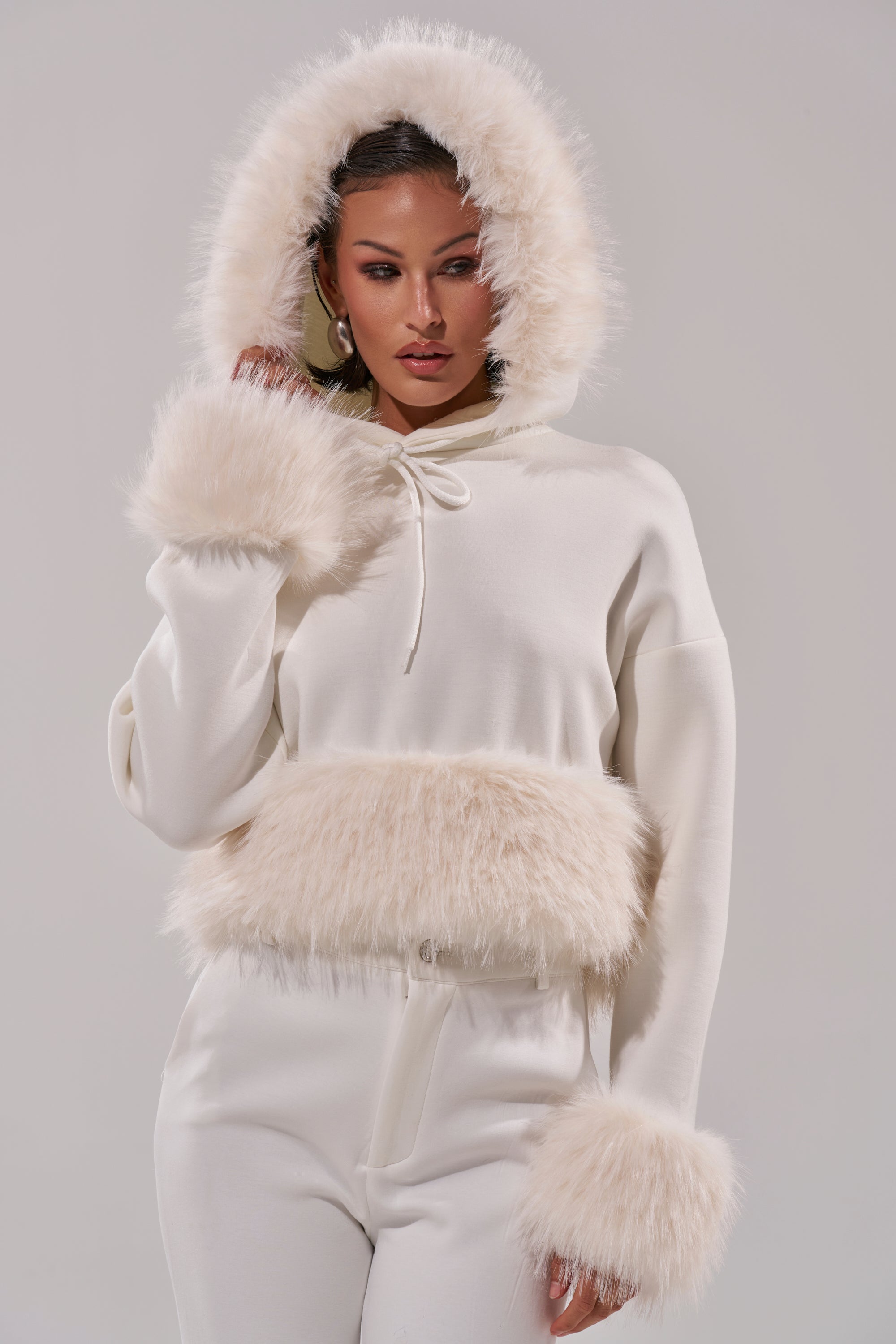 A woman poses with one hand on her hood, modeling the SNOW WHITE FUR TRIM SWEATSHIRT featuring faux fur on the hood, cuffs, and waist. The cozy top stands out against a plain light gray background. 0