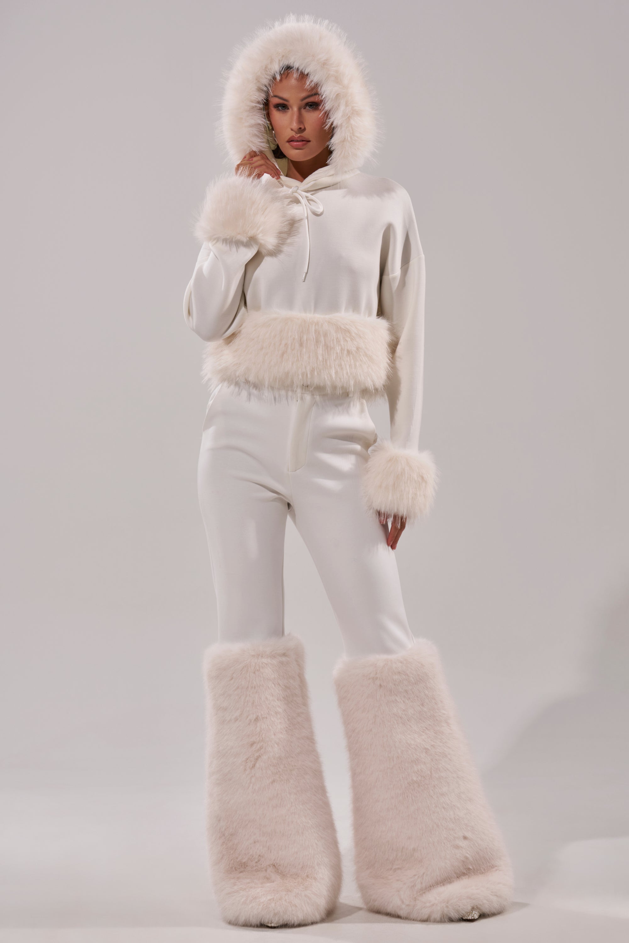 A woman models the SNOW WHITE FUR TRIM SWEATSHIRT, featuring faux fur on the hood, cuffs, and waist with oversized fluffy boot covers, against a plain background for a chic and cozy winter style. 9