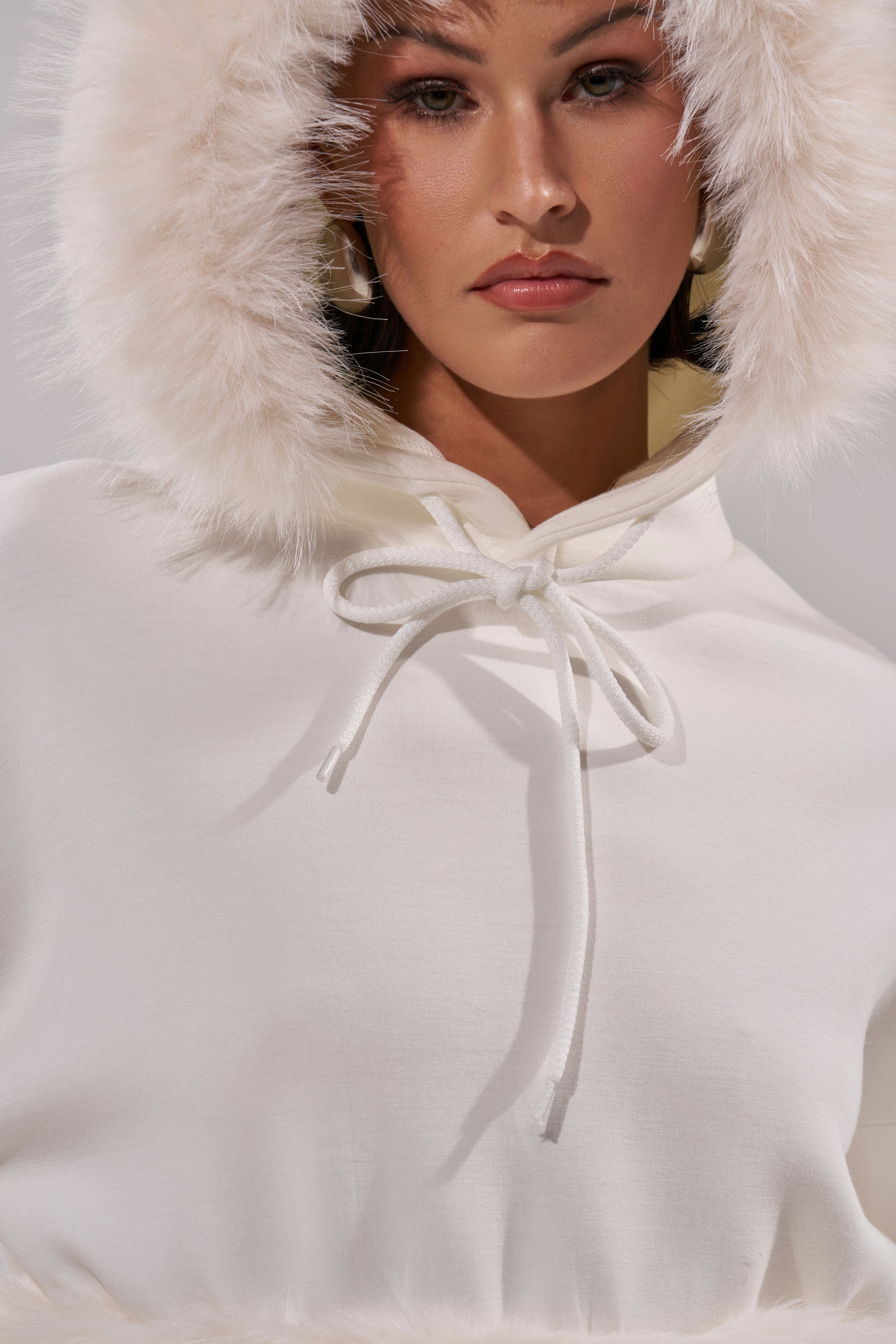 A person wears the SNOW WHITE FUR TRIM SWEATSHIRT, featuring a fluffy faux fur hood trim and tied drawstring at the neck, looking directly at the camera with the cozy hood up. 10