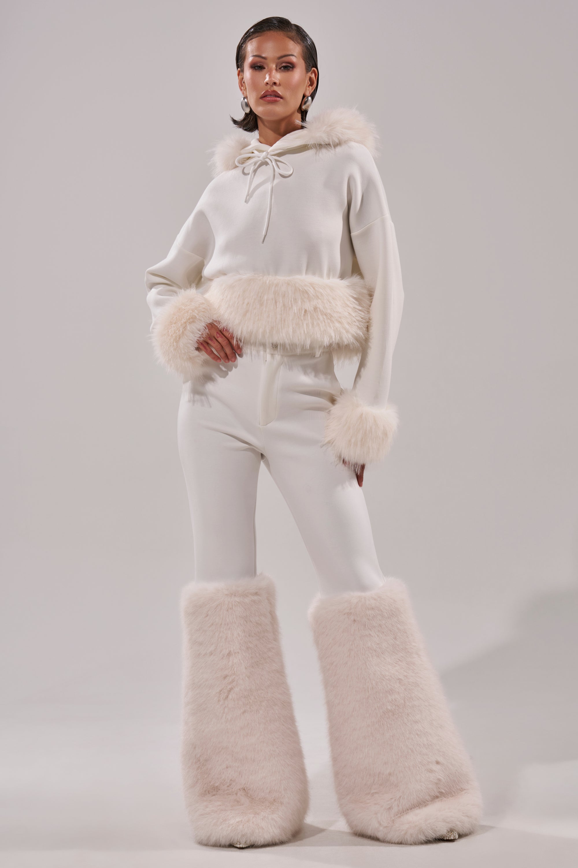 A model poses in the SNOW WHITE FUR TRIM SWEATSHIRT with faux fur on the hood, cuffs, and waist, plus oversized furry leg warmers, set against a plain light gray background. 11
