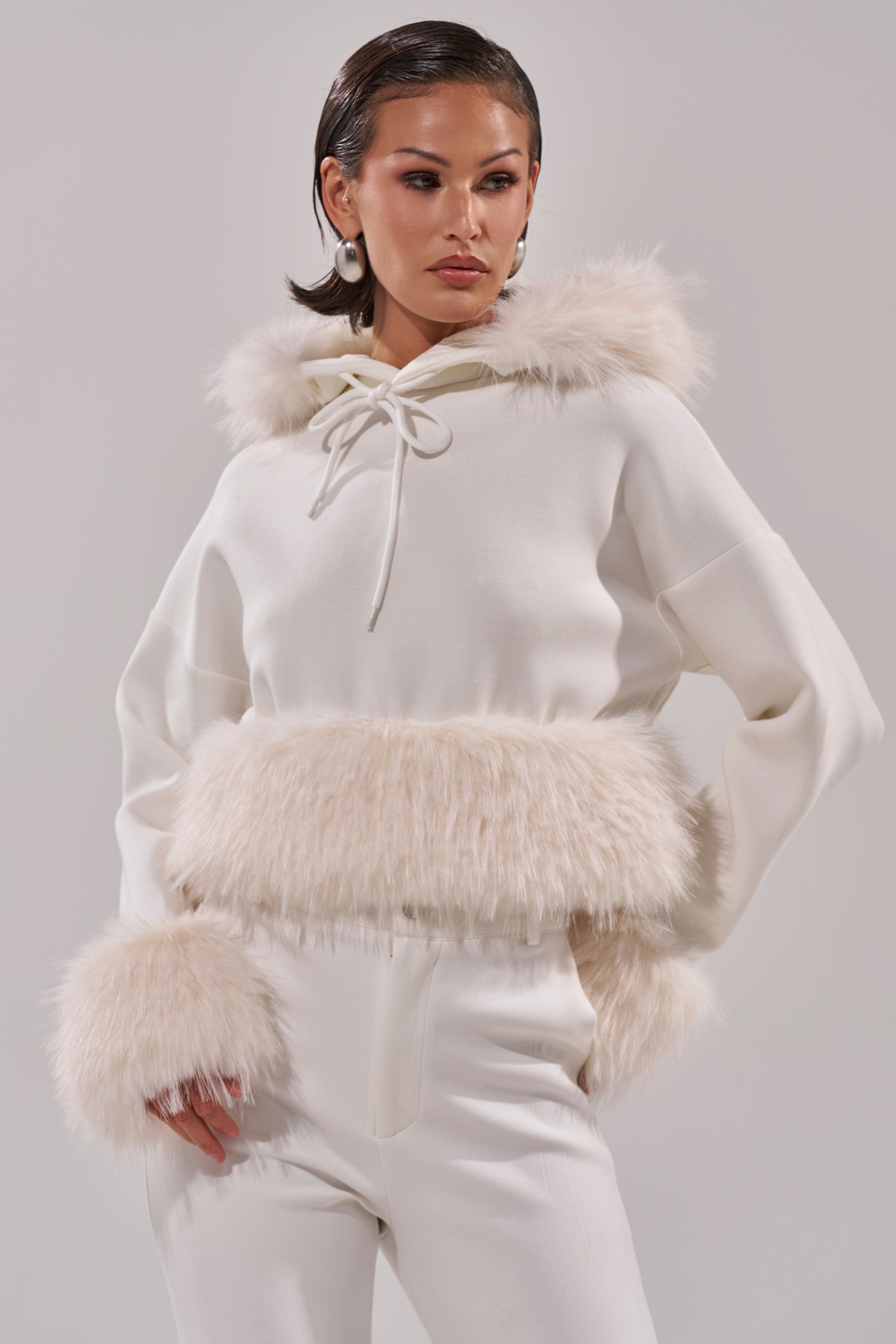 A woman models the SNOW WHITE FUR TRIM SWEATSHIRT, featuring faux fur on the hood, cuffs, and waist, paired with white high-waisted pants against a minimal light background. 1