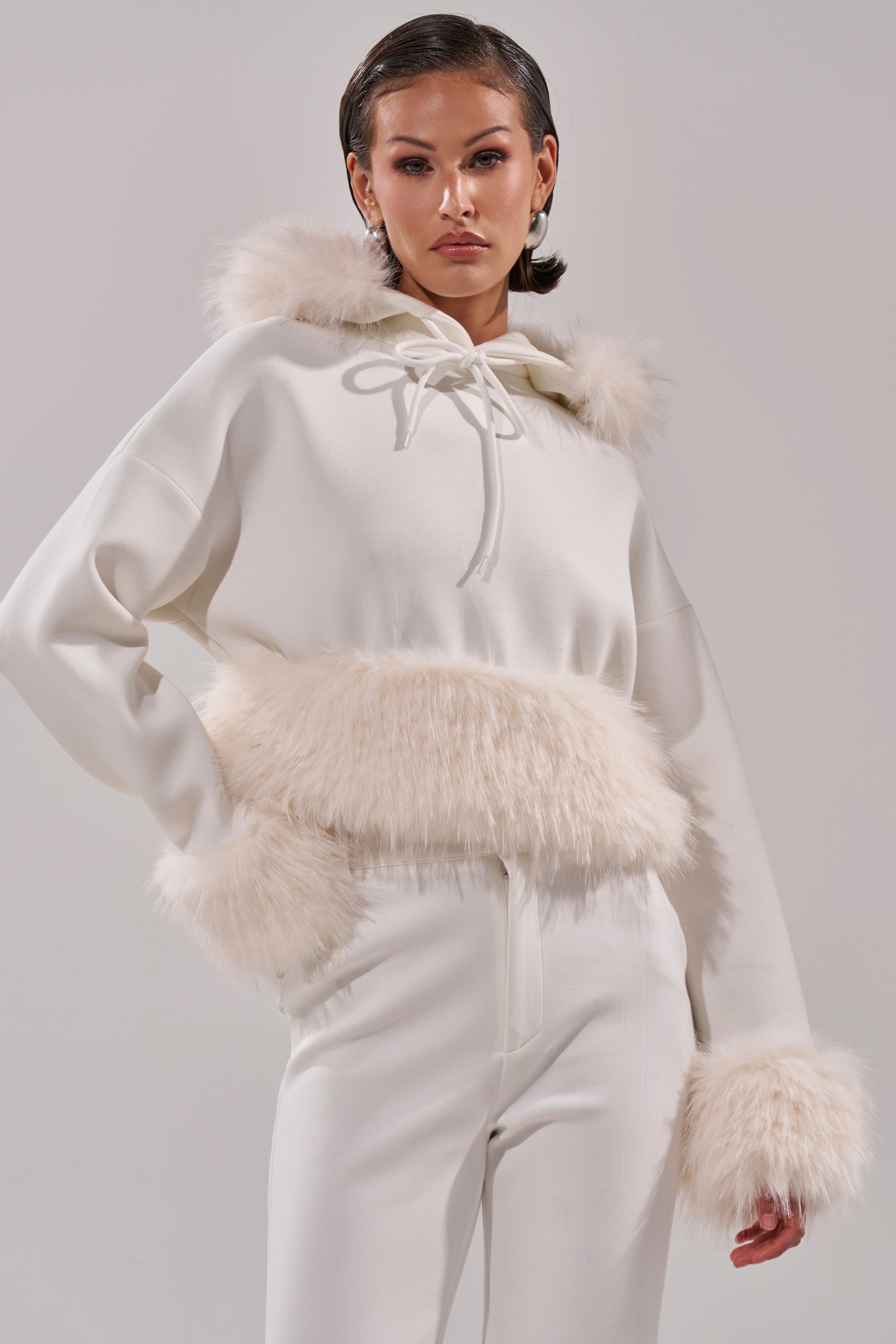 A woman models the SNOW WHITE FUR TRIM SWEATSHIRT, featuring faux fur on the hood, sleeves, and waist, styled with matching white pants against a light gray background. 2