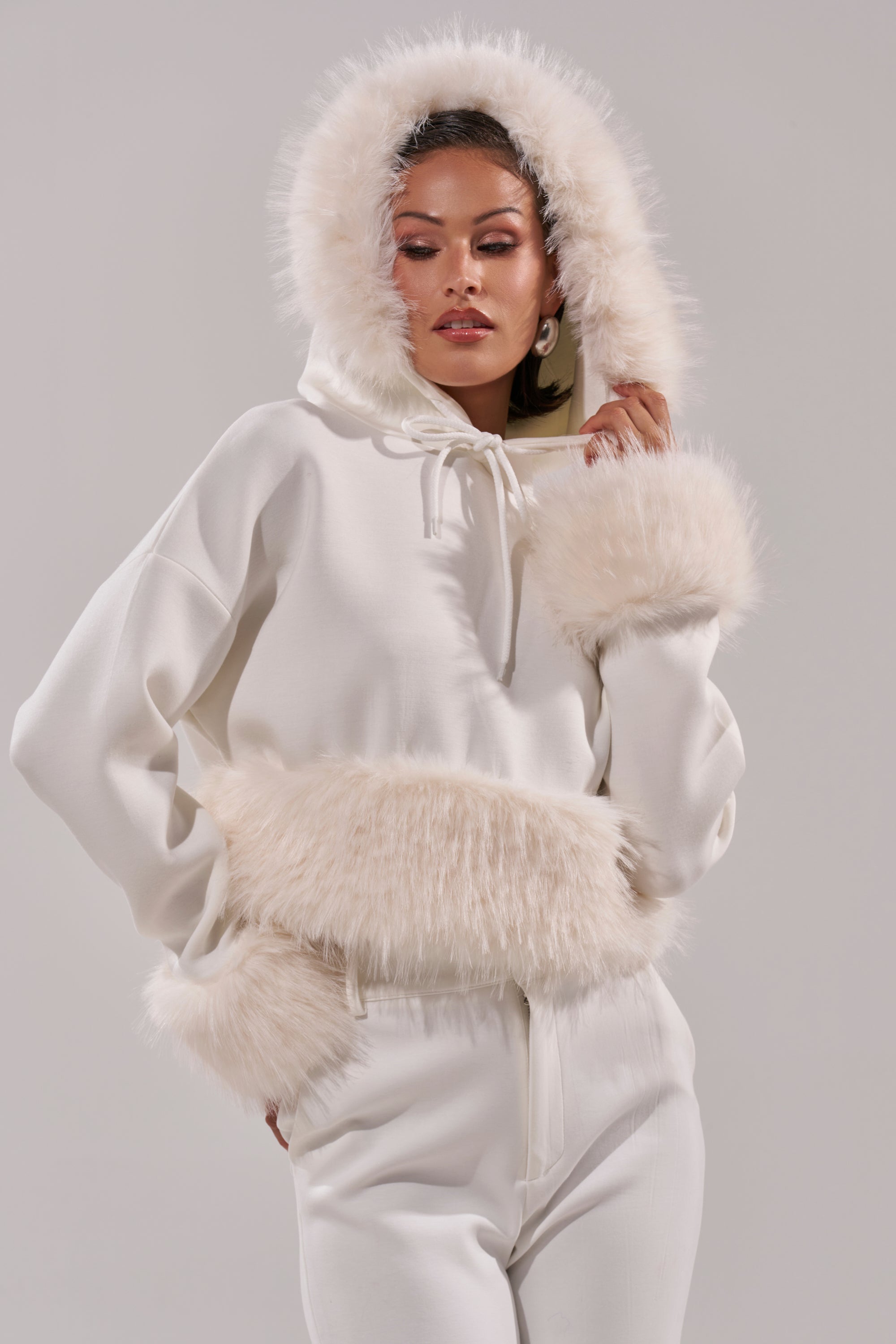 A woman models the SNOW WHITE FUR TRIM SWEATSHIRT, featuring fluffy faux fur on the hood, cuffs, and hem, with matching white pants against a neutral backdrop. 3