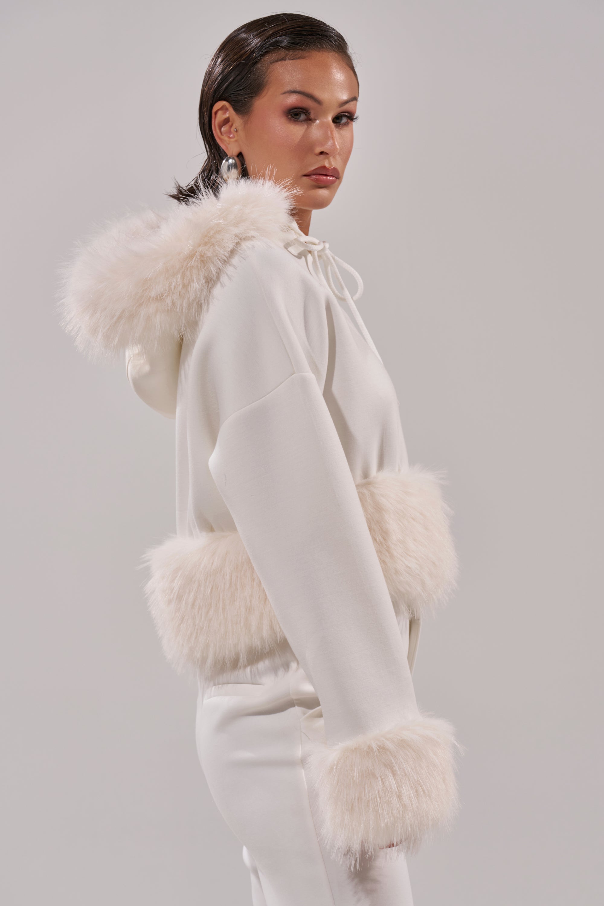 A woman wears the SNOW WHITE FUR TRIM SWEATSHIRT, featuring plush faux fur on the hood, cuffs, and waist, with matching white pants as she poses against a plain light background. 4