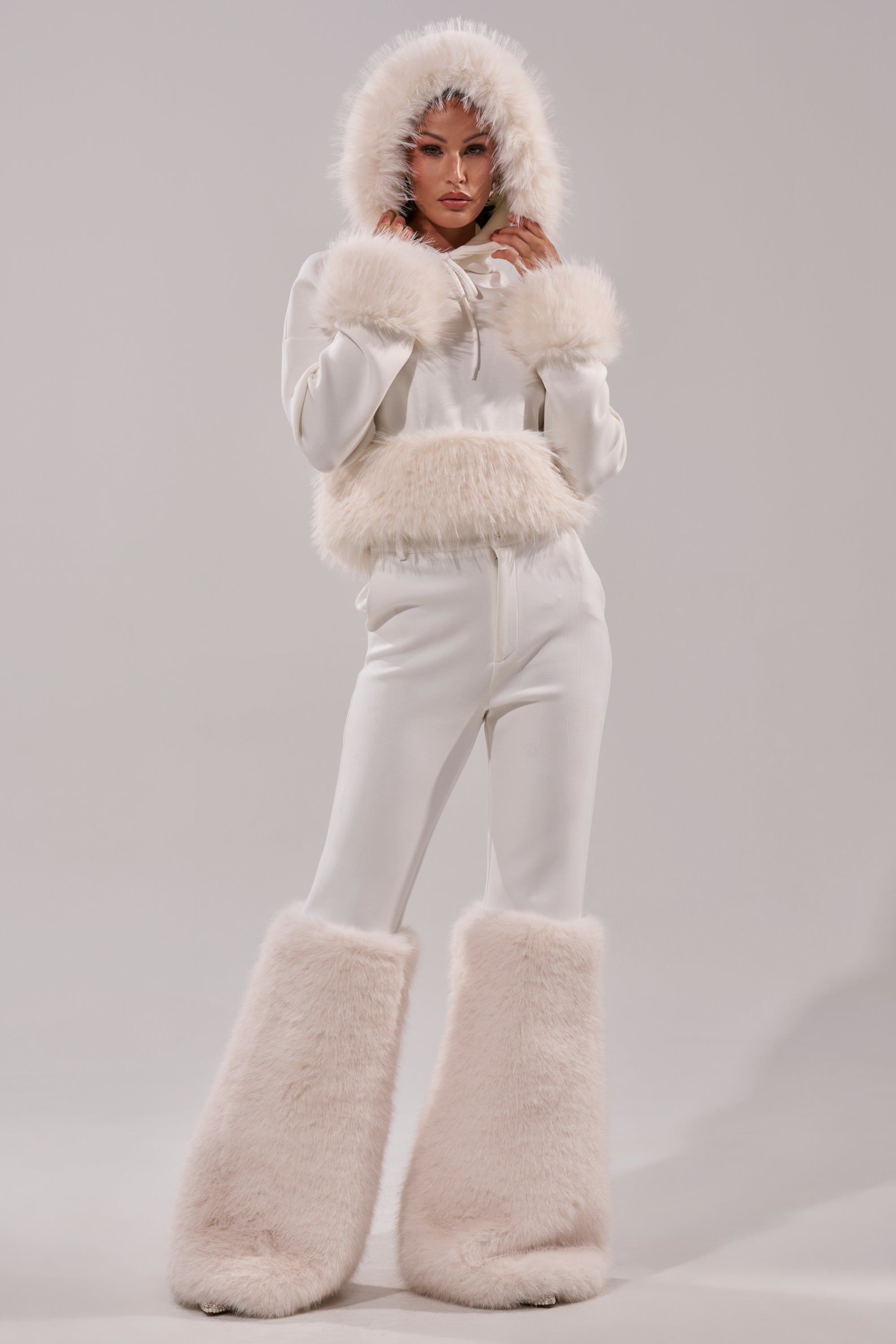 Wearing the SNOW WHITE FUR TRIM SWEATSHIRT with fluffy faux fur on the hood, cuffs, and waist, paired with oversized fur boots, a person stands against a plain light background. 6