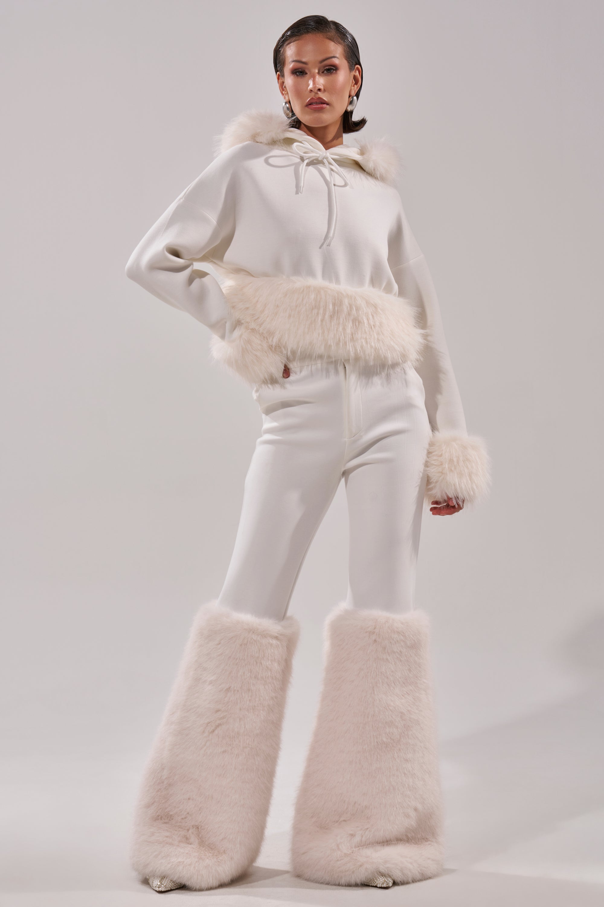 A model wearing the SNOW WHITE FUR TRIM SWEATSHIRT, featuring all-white faux fur with fluffy details on the hood, cuffs, waist, and legs, stands confidently against a plain light background for a modern, cozy look. 7