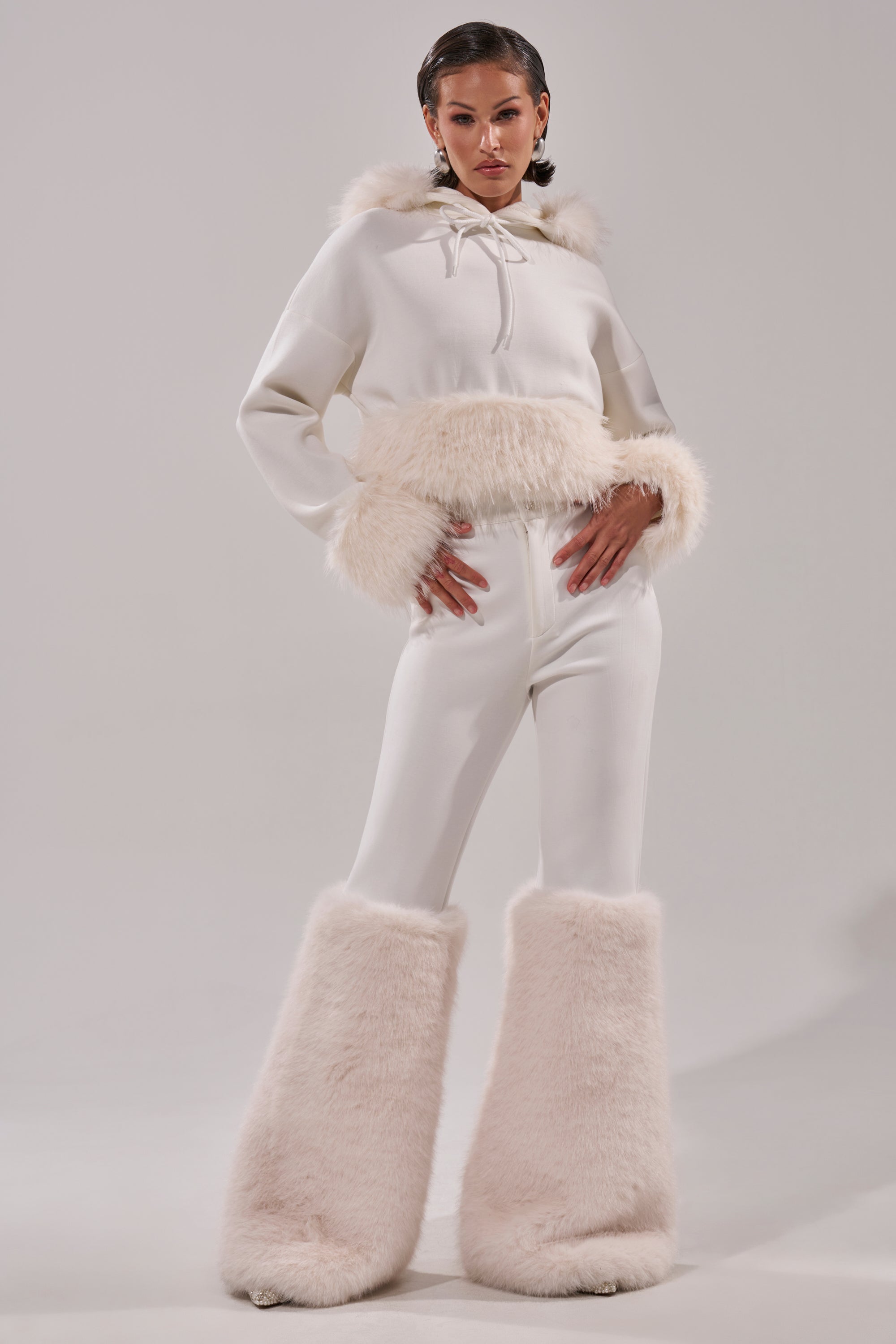 A woman poses confidently against a light gray background, wearing the SNOW WHITE FUR TRIM SWEATSHIRT—an all-white top highlighted by prominent, fluffy faux fur trim on the cuffs and hem. 8