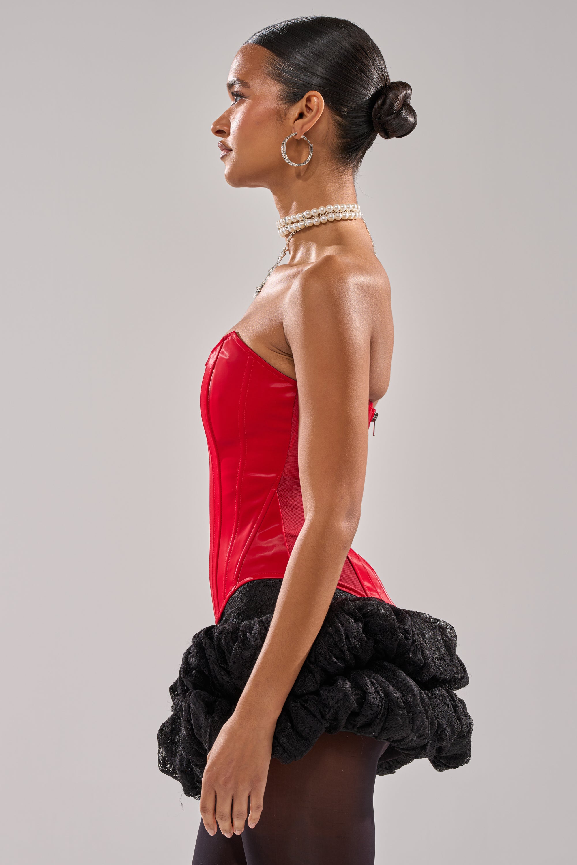 A woman poses in profile wearing the STAY OUT MY WAY CORSET IN RED, styled with a black ruffled mini skirt, sheer black tights, large hoop earrings, layered pearl necklaces, and her hair in a sleek bun. 2