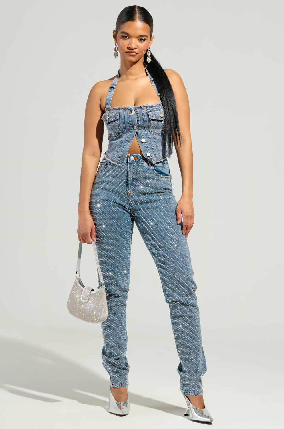 A person stands confidently in the JOLIE RHINESTONE EMBELLISHED TAPERED JEAN, paired with a denim halter top. They complete the look with circular earrings, a silver handbag, and silver heels against a plain light gray background.
