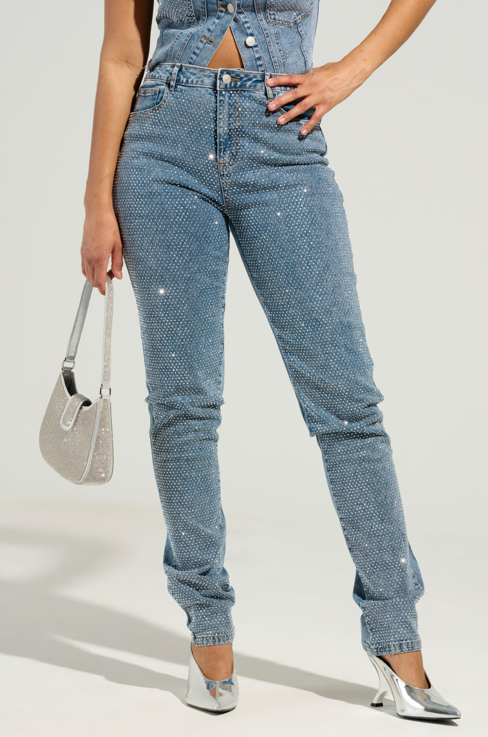 Wearing the JOLIE RHINESTONE EMBELLISHED TAPERED JEAN paired with a matching sleeveless top, a person holds a glittery purse. Their silver high-heeled shoes glimmer under the light, perfectly complementing the sparkle of their outfit. The pose is chic against a plain background.