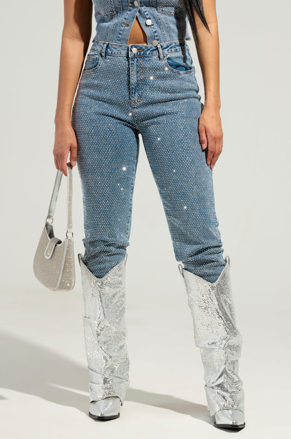 A woman dons the Jolie Rhinestone Embellished Tapered Jeans, paired with dazzling silver sequin boots and a chic small silver handbag. She stands confidently against a plain background.