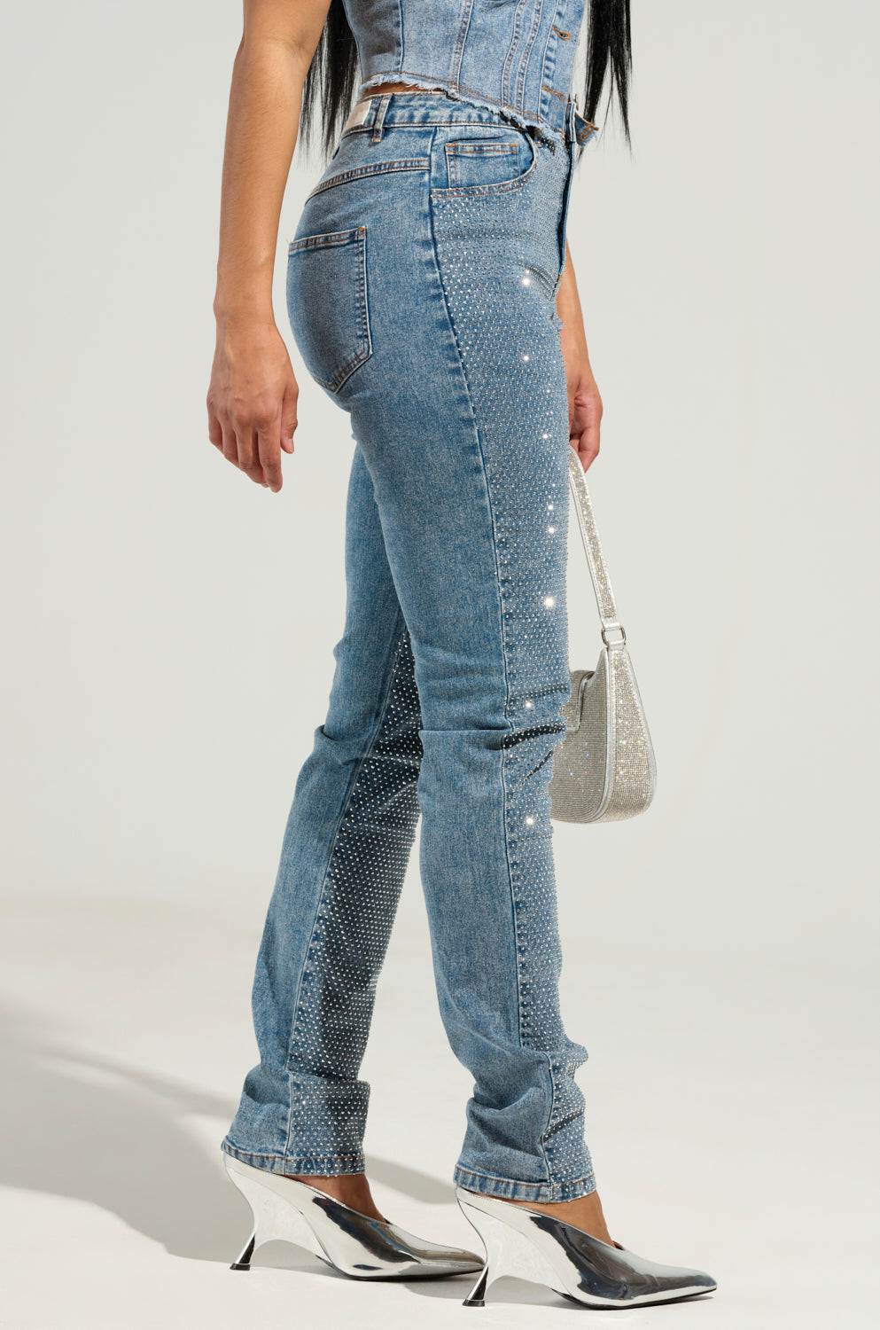 A person dressed in JOLIE RHINESTONE EMBELLISHED TAPERED JEANS, featuring shimmering embellishments along the sides and hems, is holding a silver purse and wearing metallic wedge heels against a plain, light-colored background.