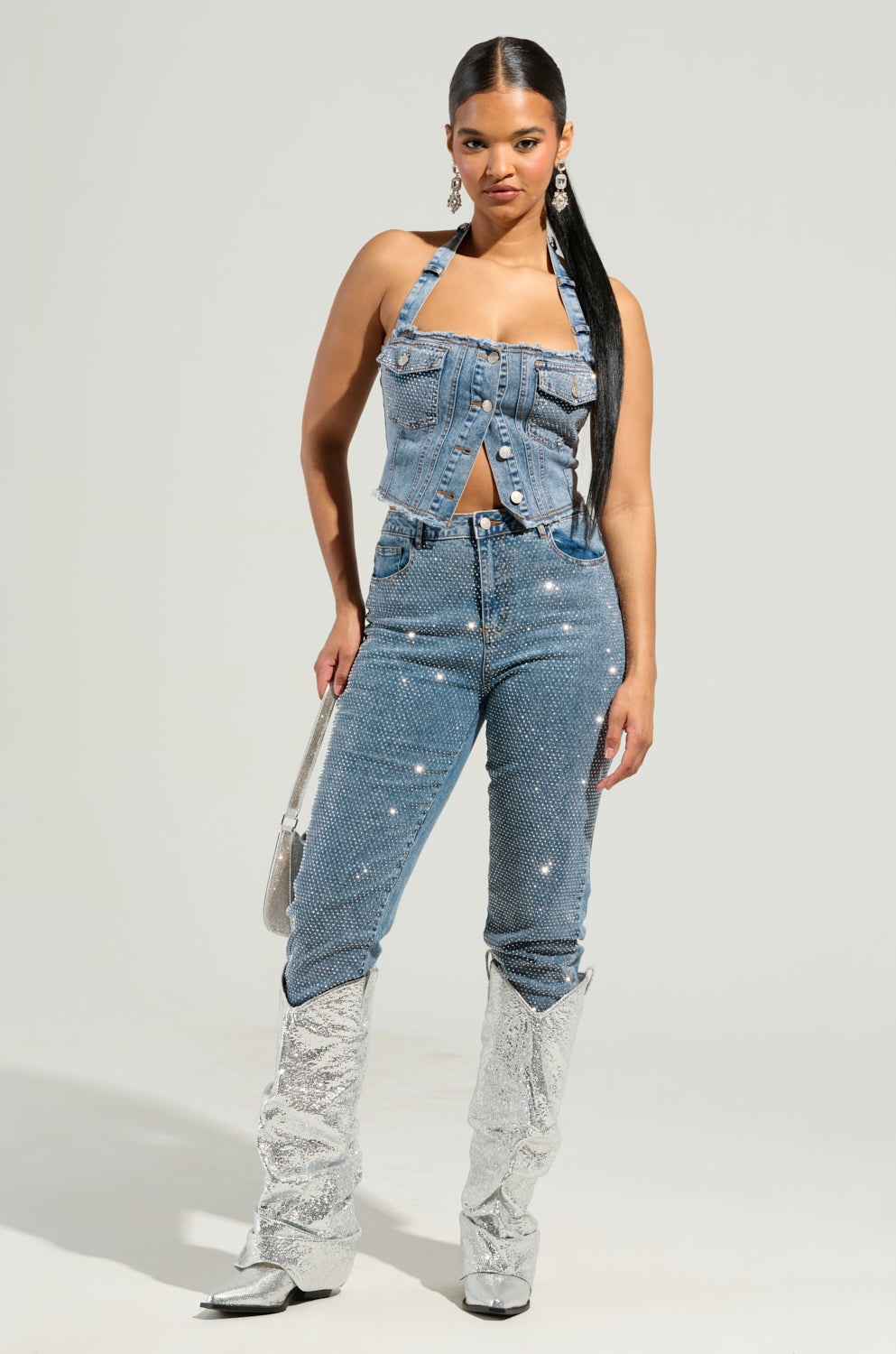A woman showcases the Jolie Rhinestone Embellished Tapered Jeans, paired with a halter top adorned in shimmering silver rhinestones. Her outfit is polished off with silver sequined high-heeled boots and a stylish silver handbag. Her sleek long hair beautifully complements her statement earrings.