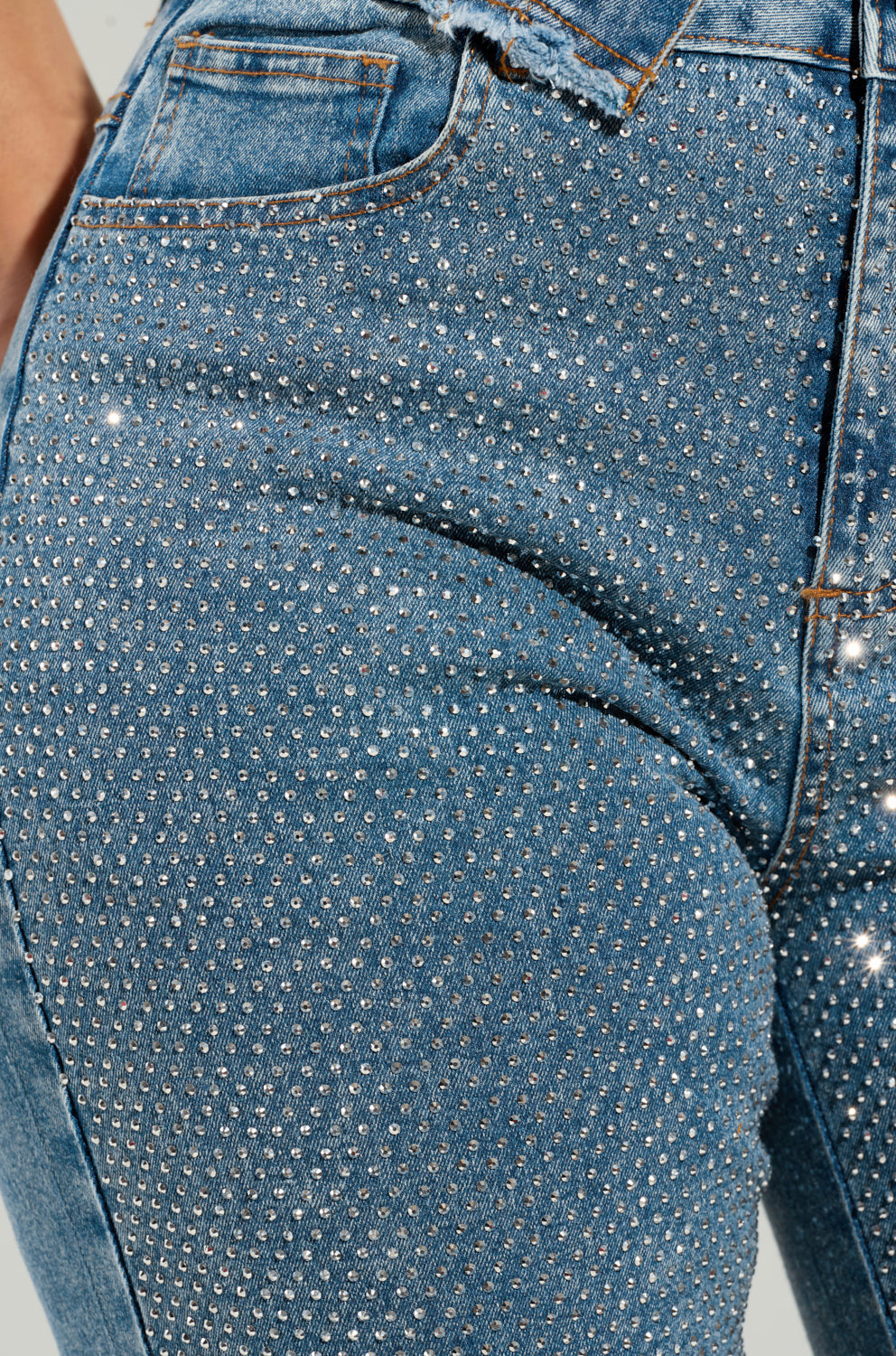 Introducing the JOLIE RHINESTONE EMBELLISHED TAPERED JEAN, showcasing a close-up of high-rise denim adorned with small, sparkling silver rhinestones. These jeans feature a frayed edge at the waistband and intricate stitching details around the pockets, producing a shimmering effect across their surface.
