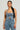 A person with long black hair stands confidently, dressed in the JOLIE RHINESTONE EMBELLISHED DENIM CORSET paired with matching pants. The outfit features delicate distressed detailing and is accessorized with hoop earrings. They pose against a plain background, emphasizing their striking silhouette.