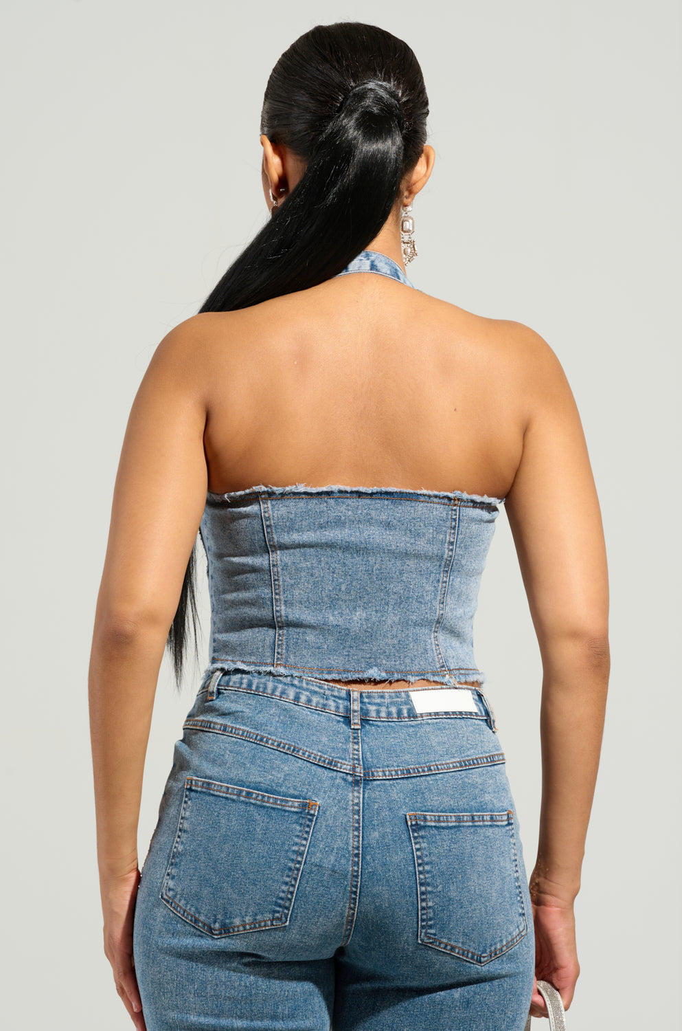 A rear view of a person with long black hair in a ponytail, showcasing the Jolie Collection's rhinestone embellished denim corset and jeans. The sleeveless corset complements the jeans with back pockets, set against a neutral background.