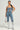 A person stands confidently, wearing the JOLIE RHINESTONE EMBELLISHED DENIM CORSET paired with matching jeans adorned with sequins. They have long, straight hair and hold a small white purse. The outfit is completed with shiny, silver knee-high boots.